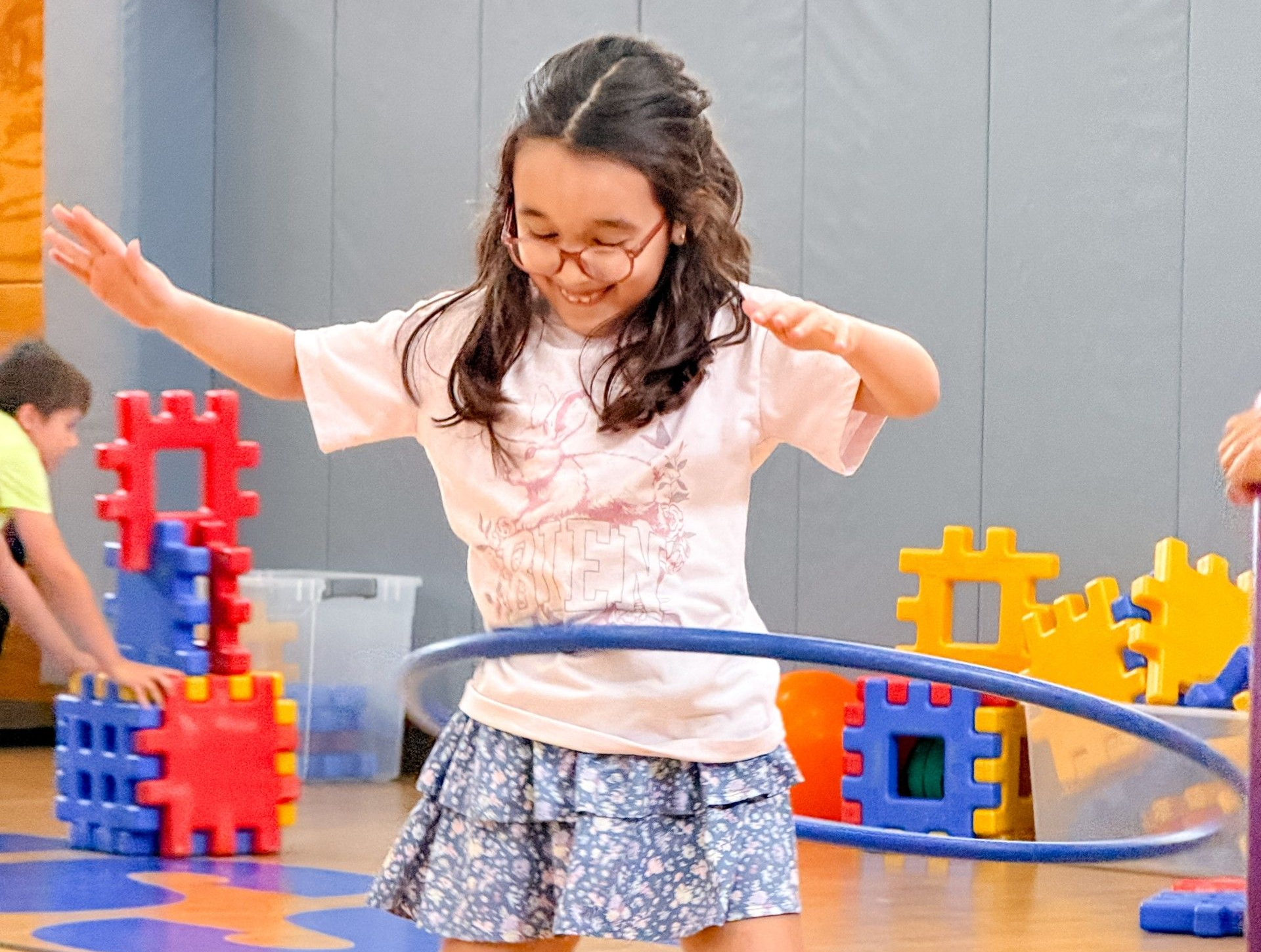 A girl in a pink dress in a gymnasium hunched over a green hula-hoop.