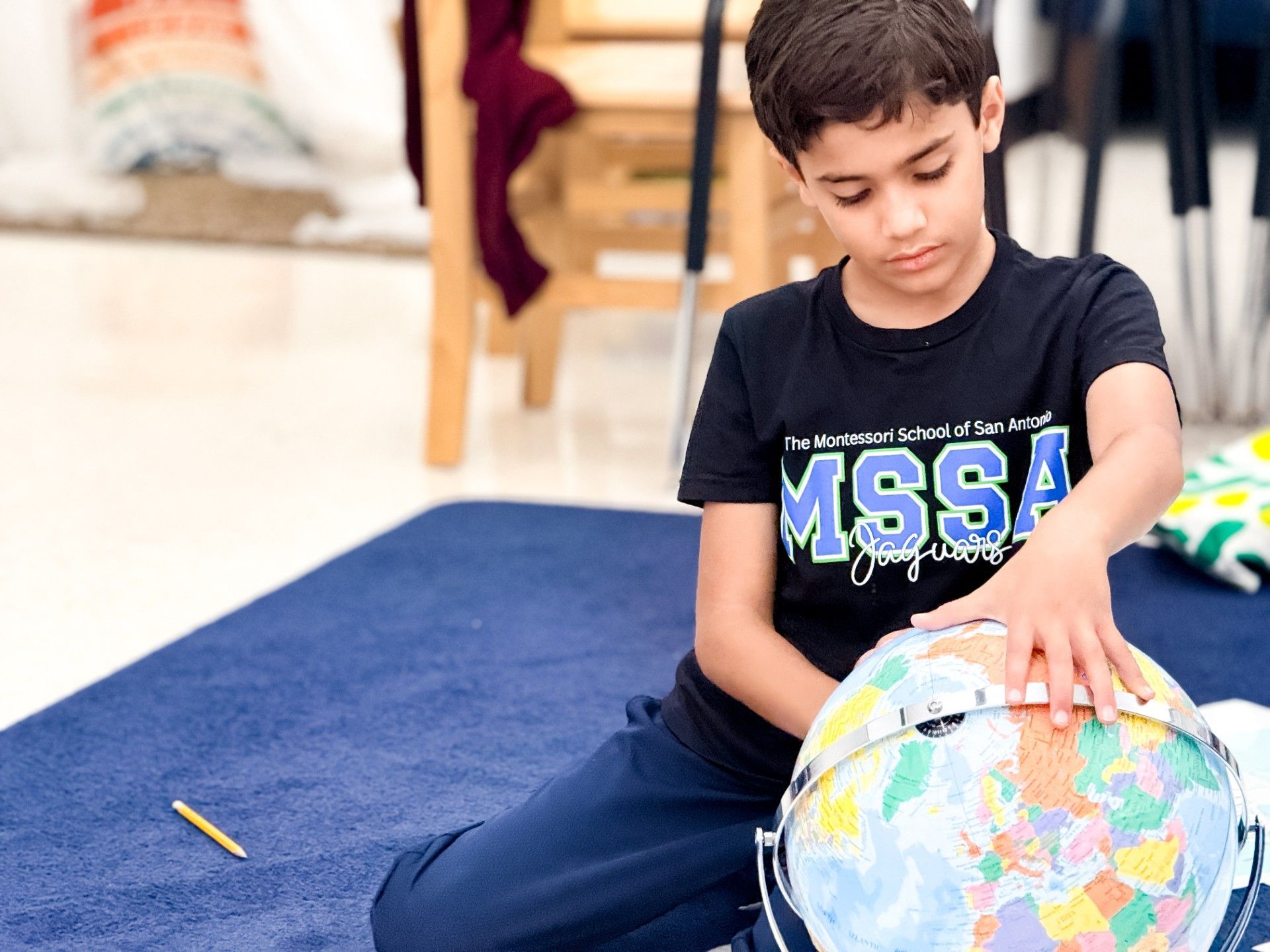 Boy sitting on the floor beside a globe, pointing at it in a classroom setting