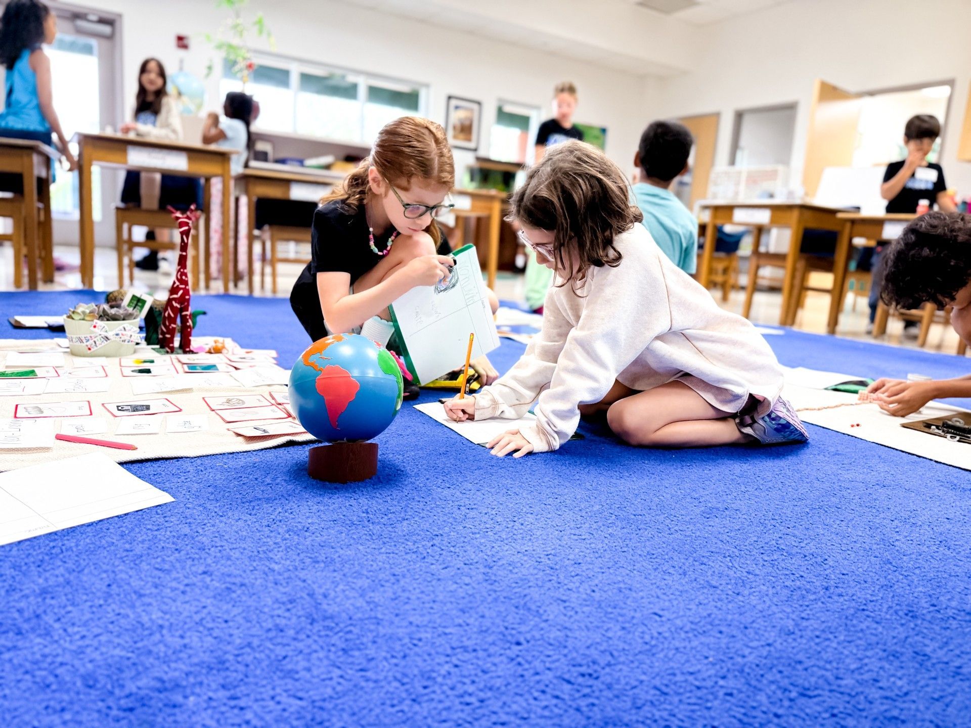 Children working on a floor activity in a classroom, with a globe and scattered papers on blue carpet.