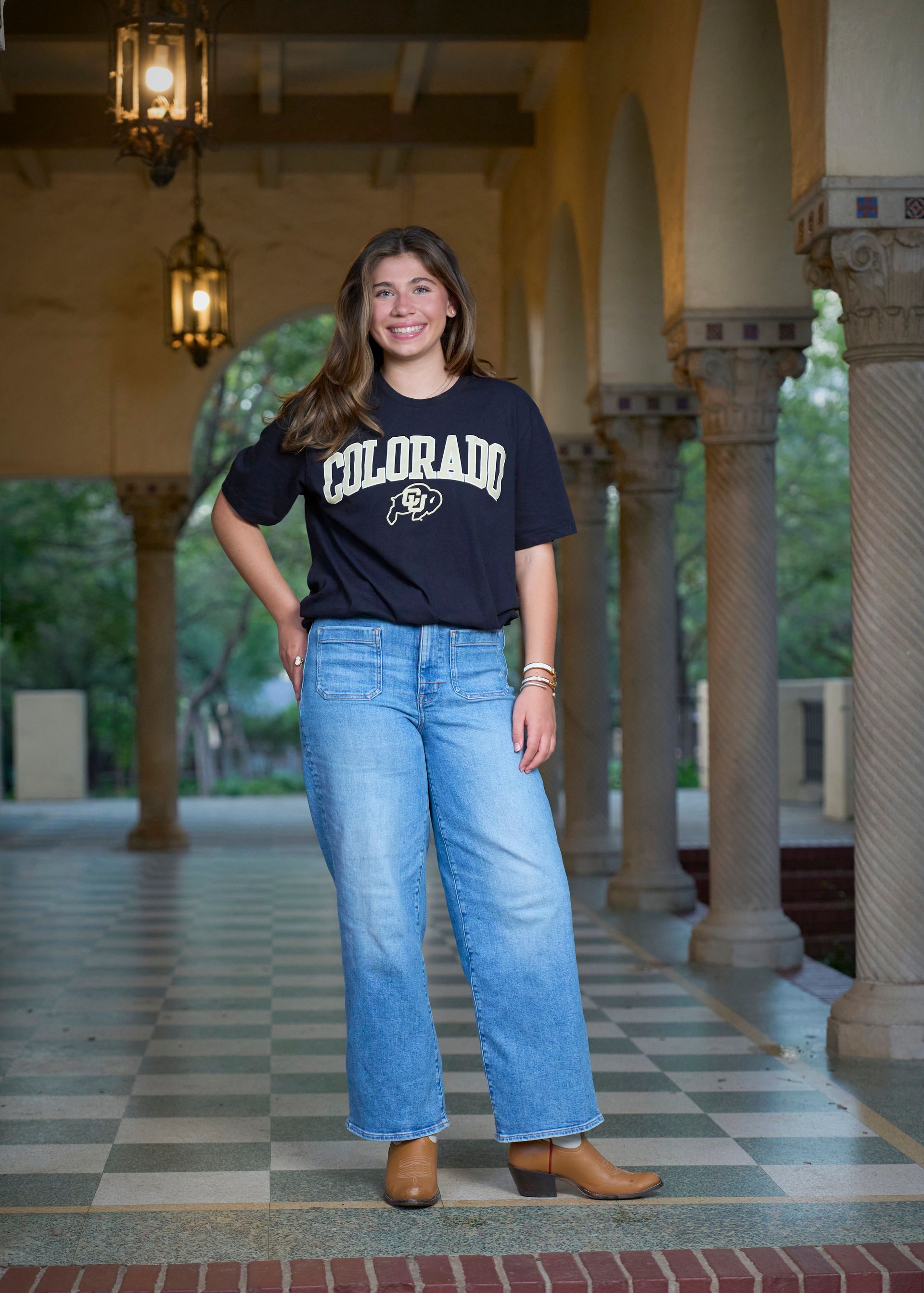 Girl Standing in a hallway of columns with a University of Colorado shirt on and jeans.