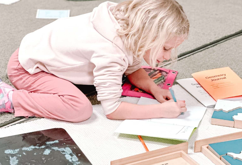 Girl in pink sweatsuit and hoodie drawing on the floor with books and wooden puzzles around her.
