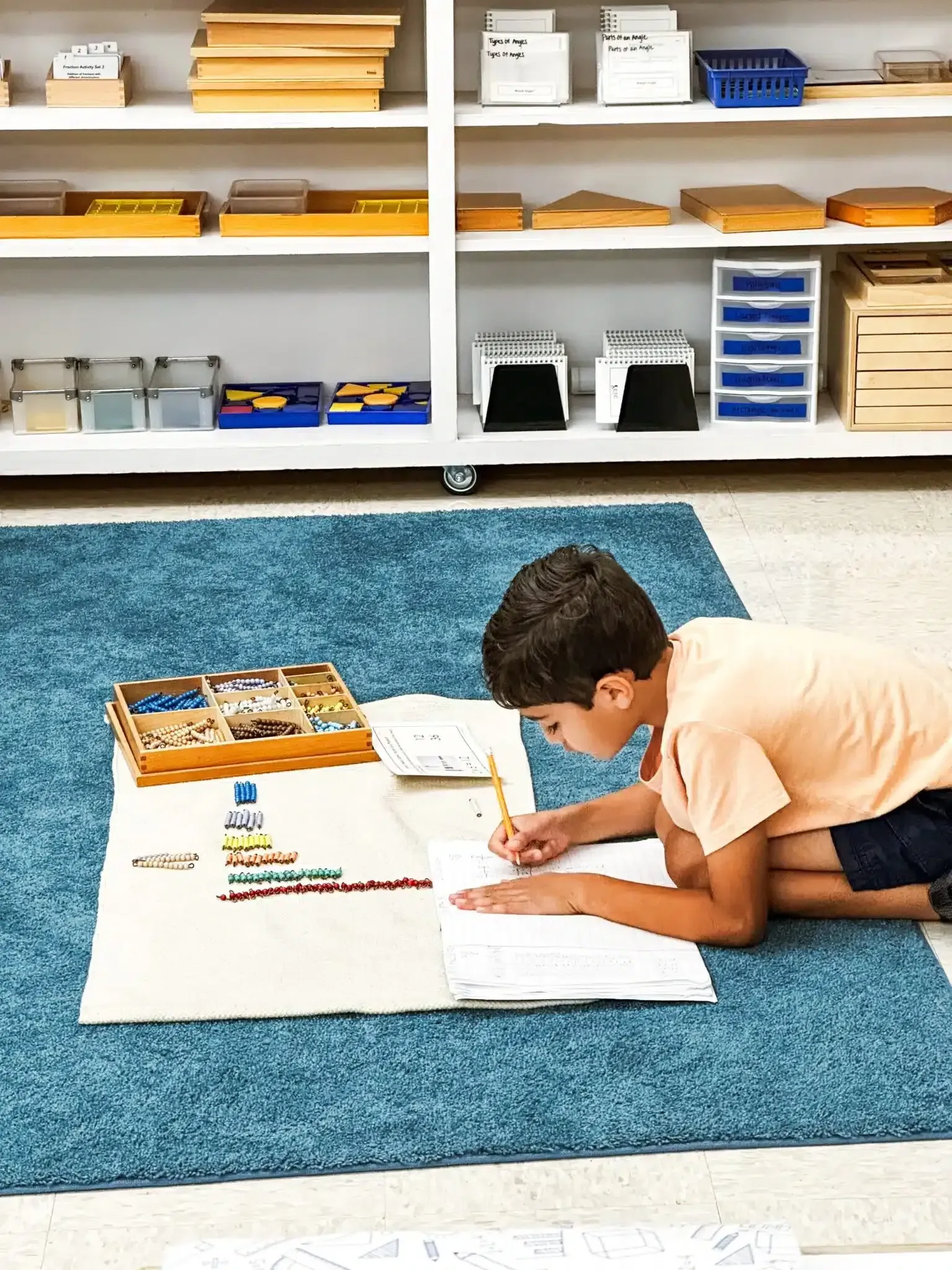 Boy writing on paper on a blue rug, surrounded by Montessori materials in a classroom setting.