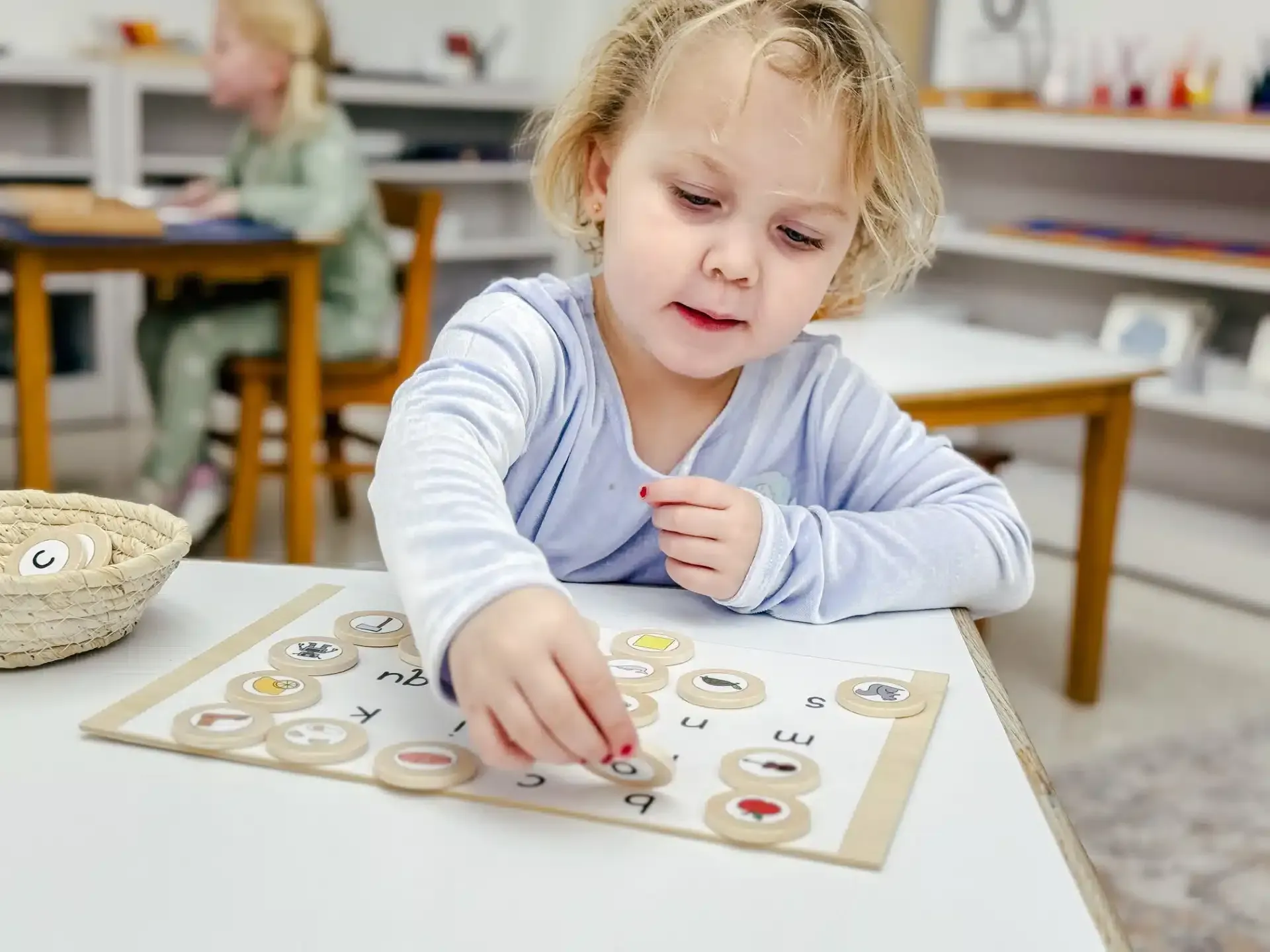 Young child at a table playing with a matching game, focused expression. Classroom setting with another child in background.