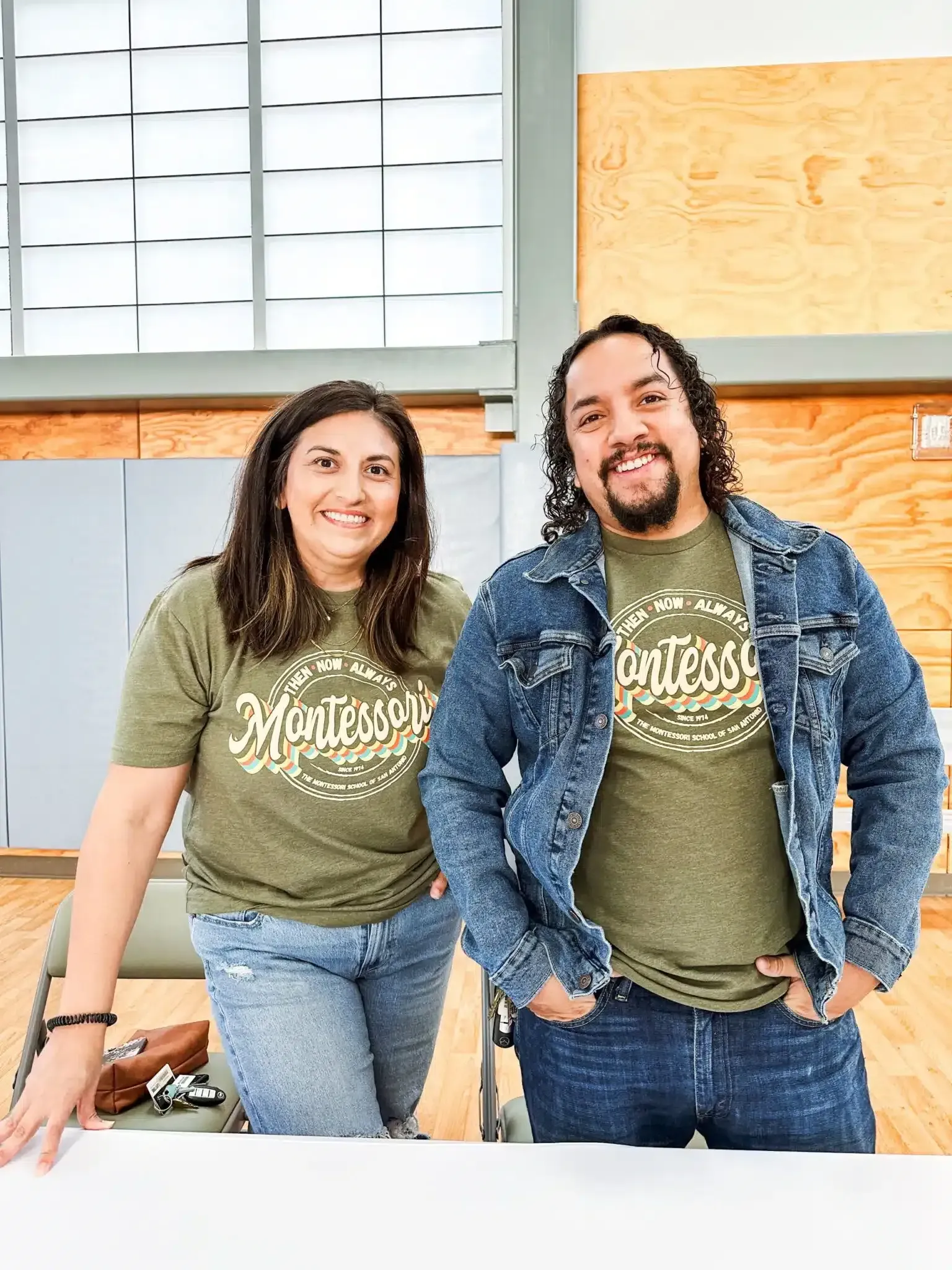 Two people wearing matching green shirts with logo, smiling. Man in denim jacket. Interior shot.