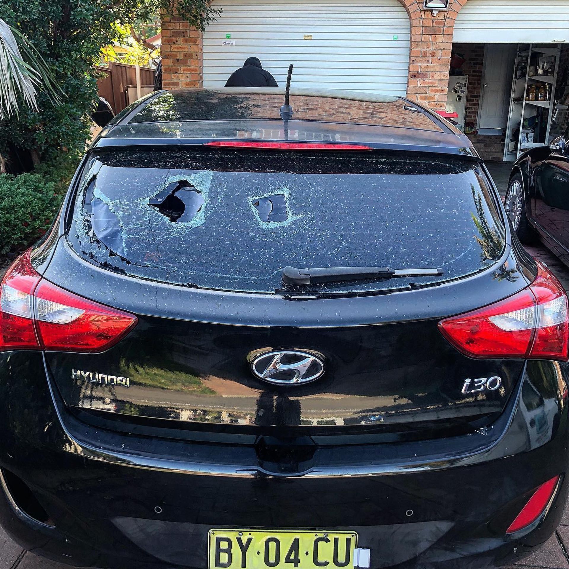 Black Hyundai hatchback with shattered rear window and visible damage, parked in a driveway.