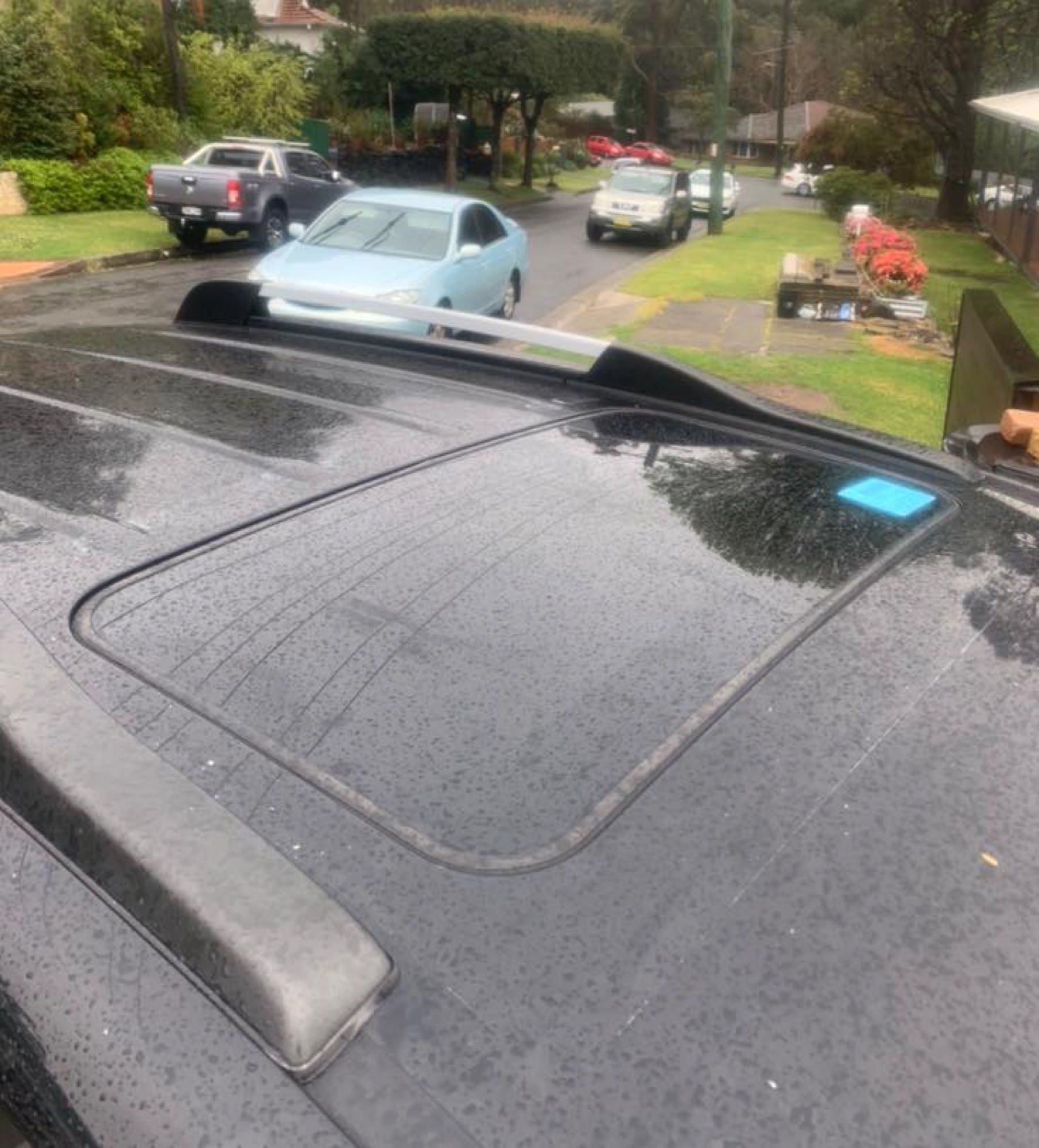 Rain-speckled car roof overlooking a residential street with parked cars and wet greenery