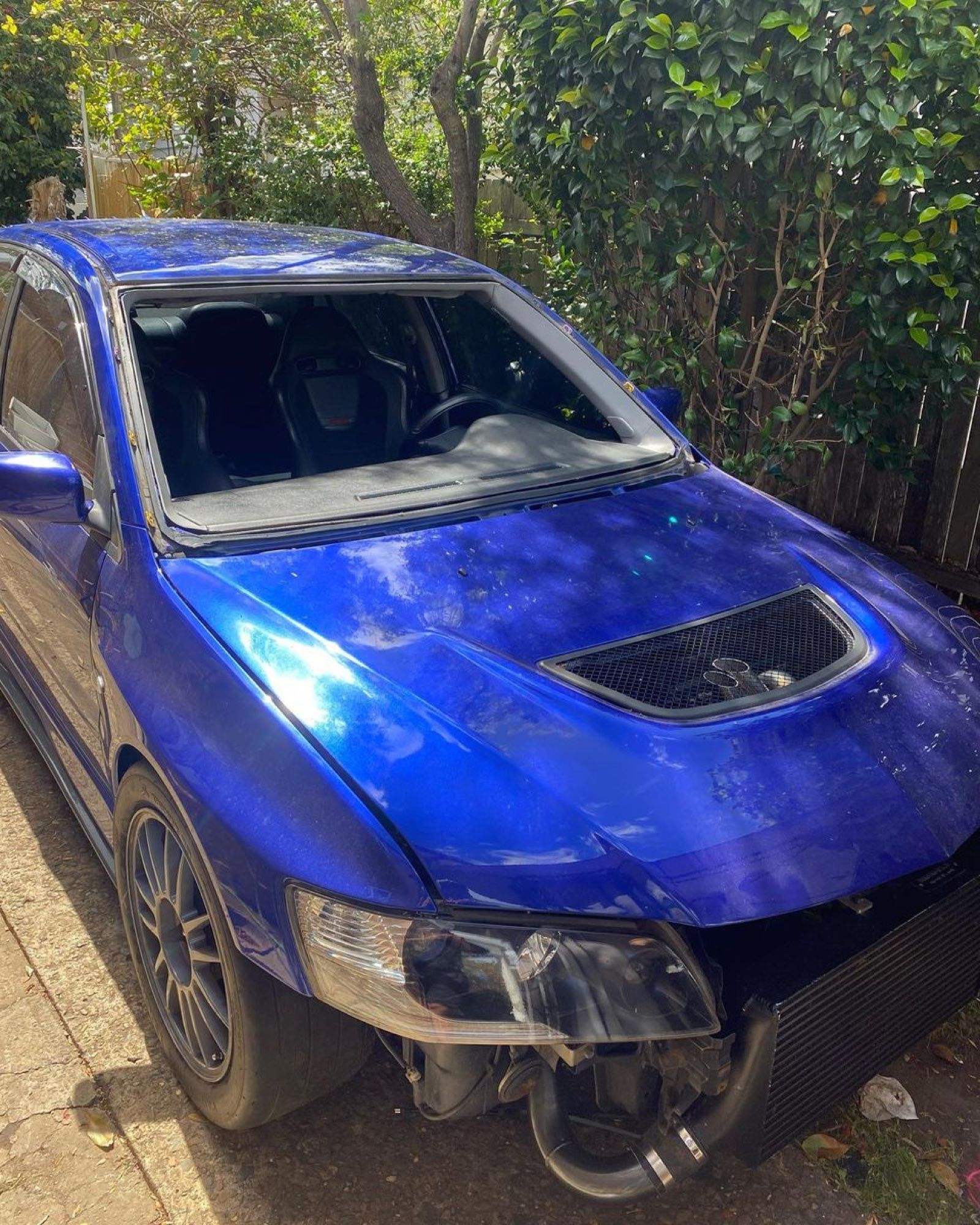 Blue car with a shattered windshield and damaged front bumper parked by bushes
