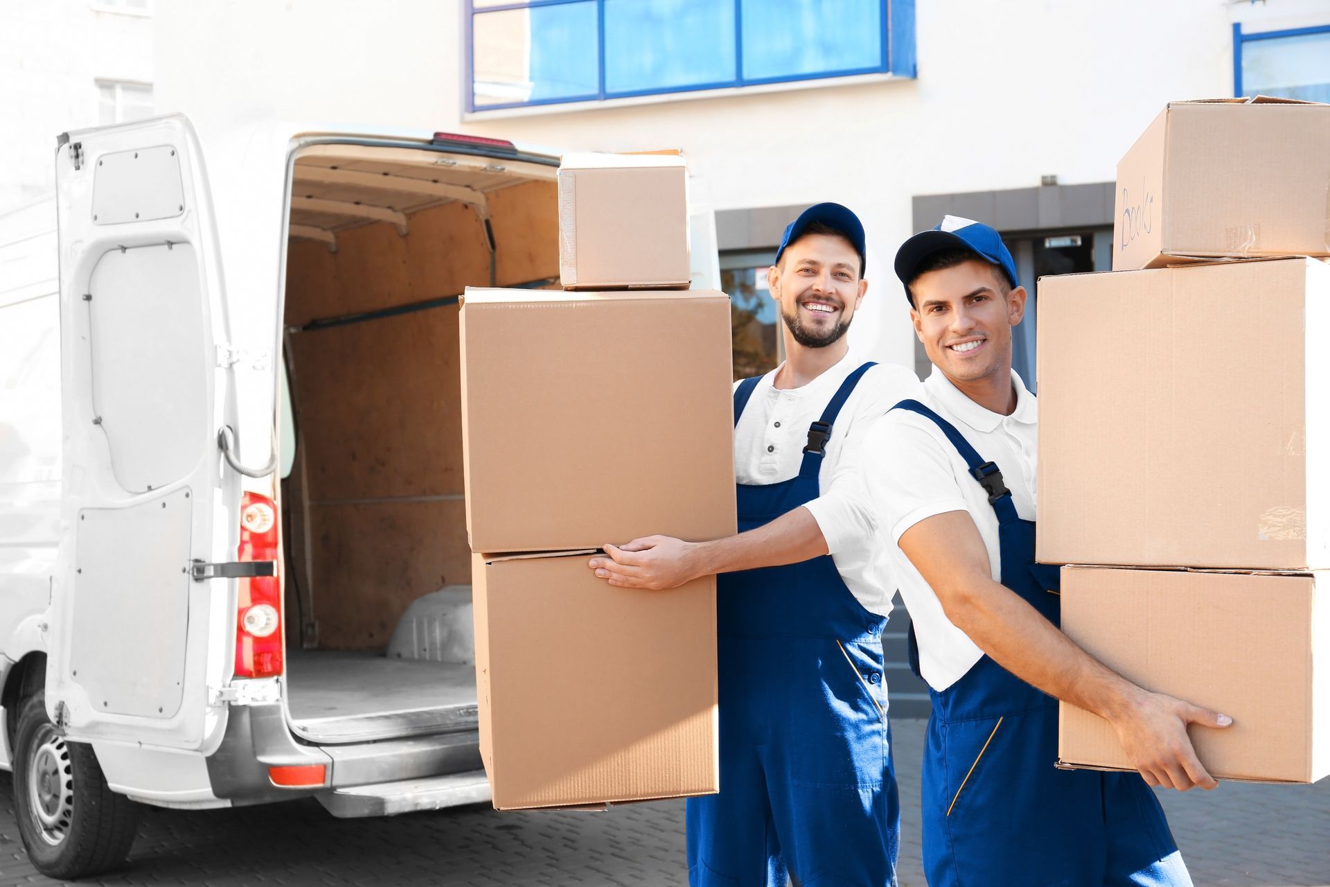Two workers in blue overalls carry stacked boxes by an open delivery van.