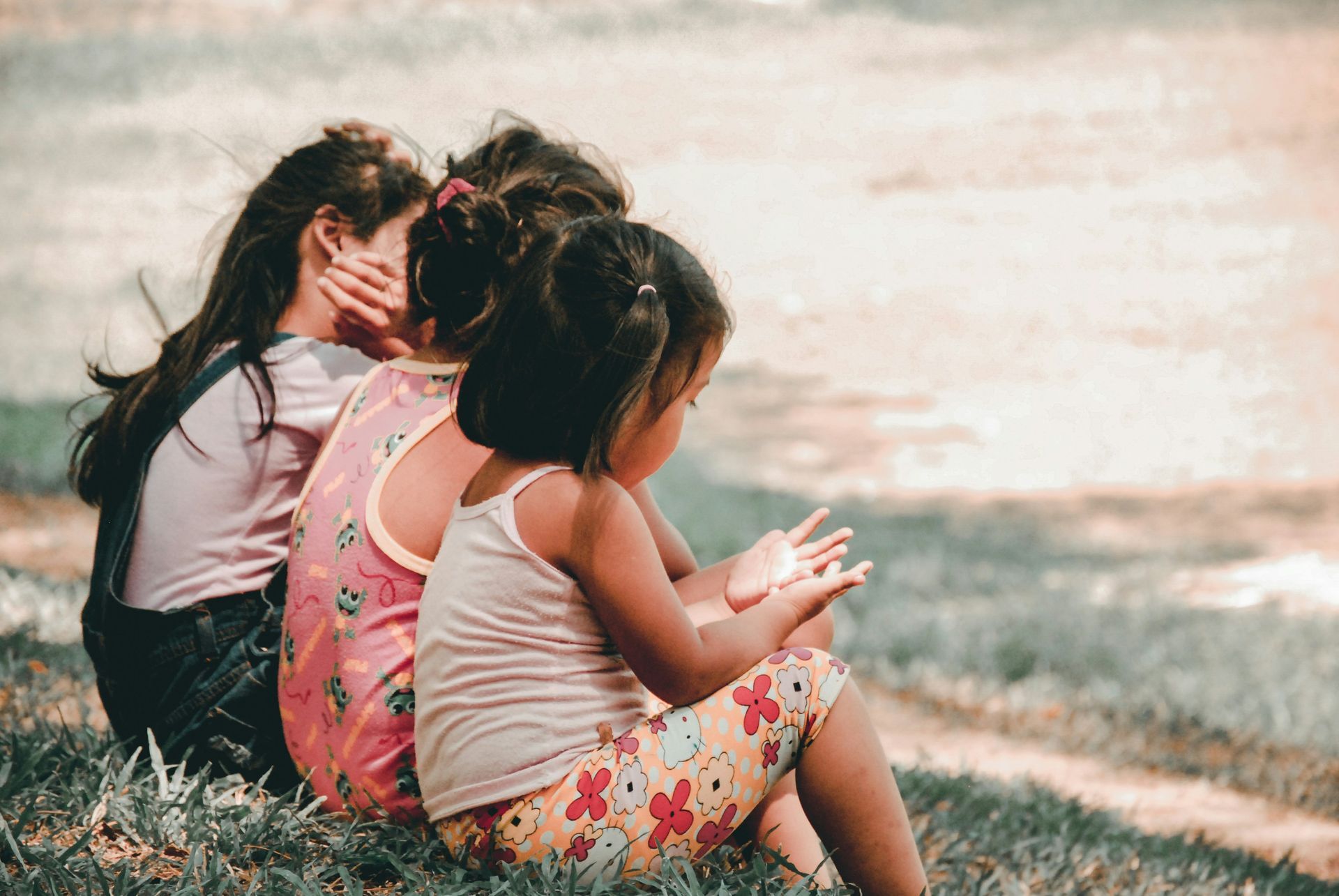 Three little girls are sitting on the grass near the water.