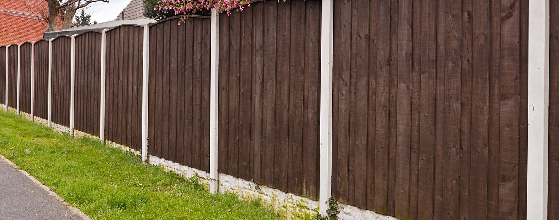A brown wooden fence with white posts is next to a grassy sidewalk.