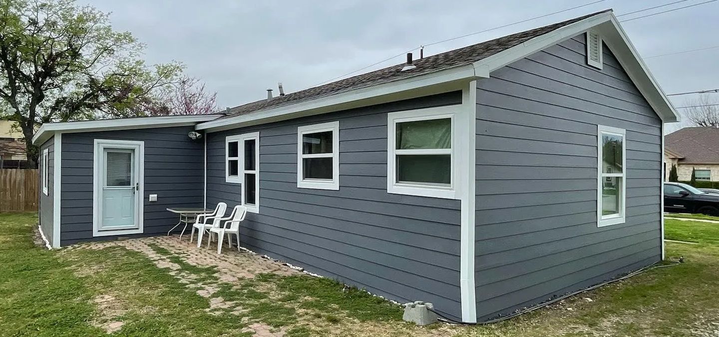 A small gray house with white trim is sitting on top of a lush green field.