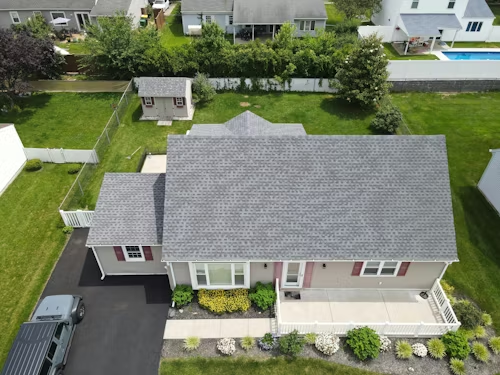 Aerial view of a gray-roofed suburban house with driveway, front porch, and fenced backyard.