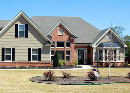 Two-story house with tan siding, brick facade, black shutters, and well-manicured lawn under a blue sky.