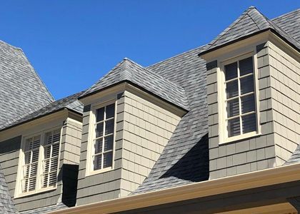 Three dormer windows with gray shingles and beige siding against a blue sky.