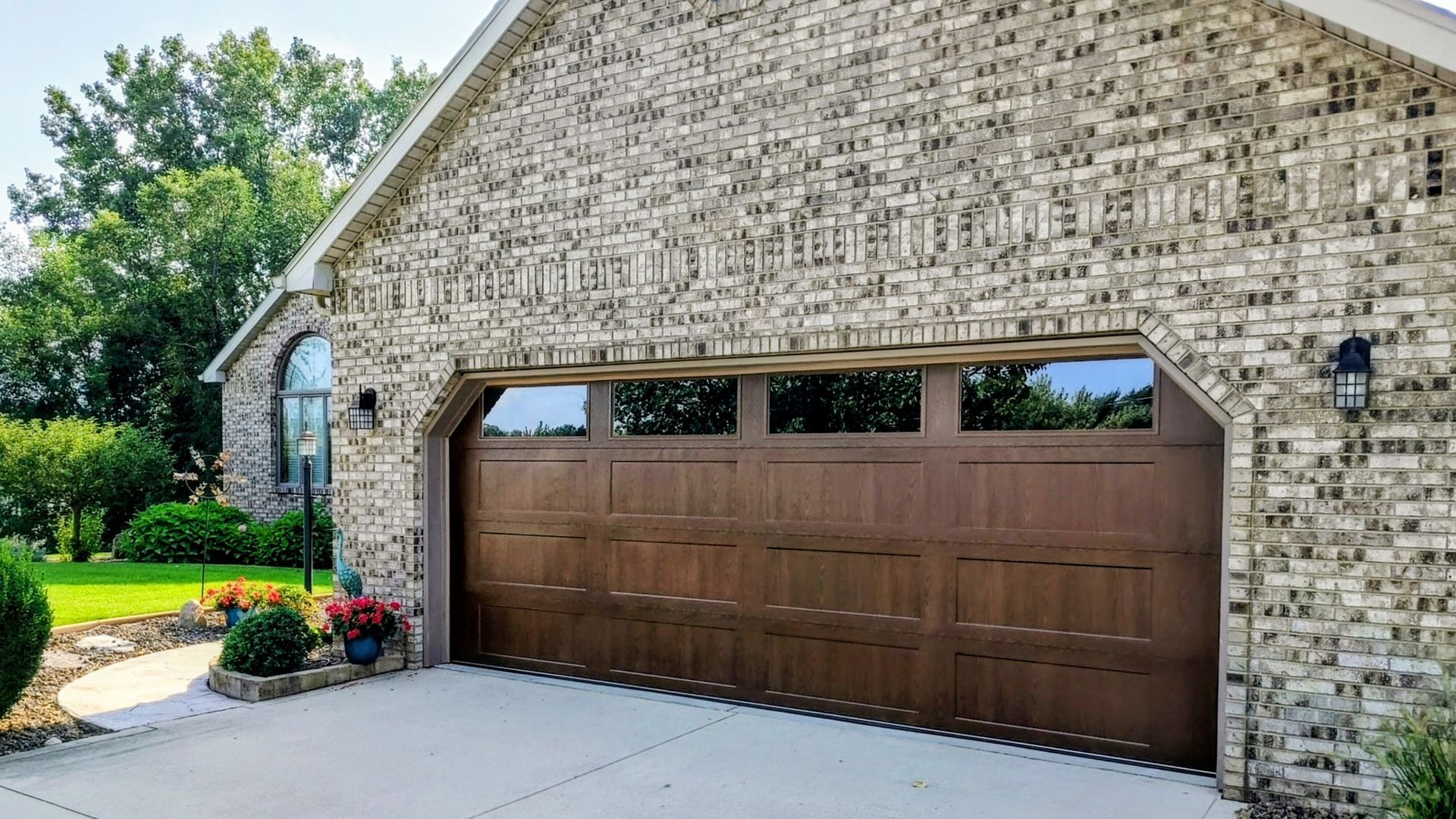 Brick house exterior with a brown garage door, driveway, and flowerbed under bright sunlight