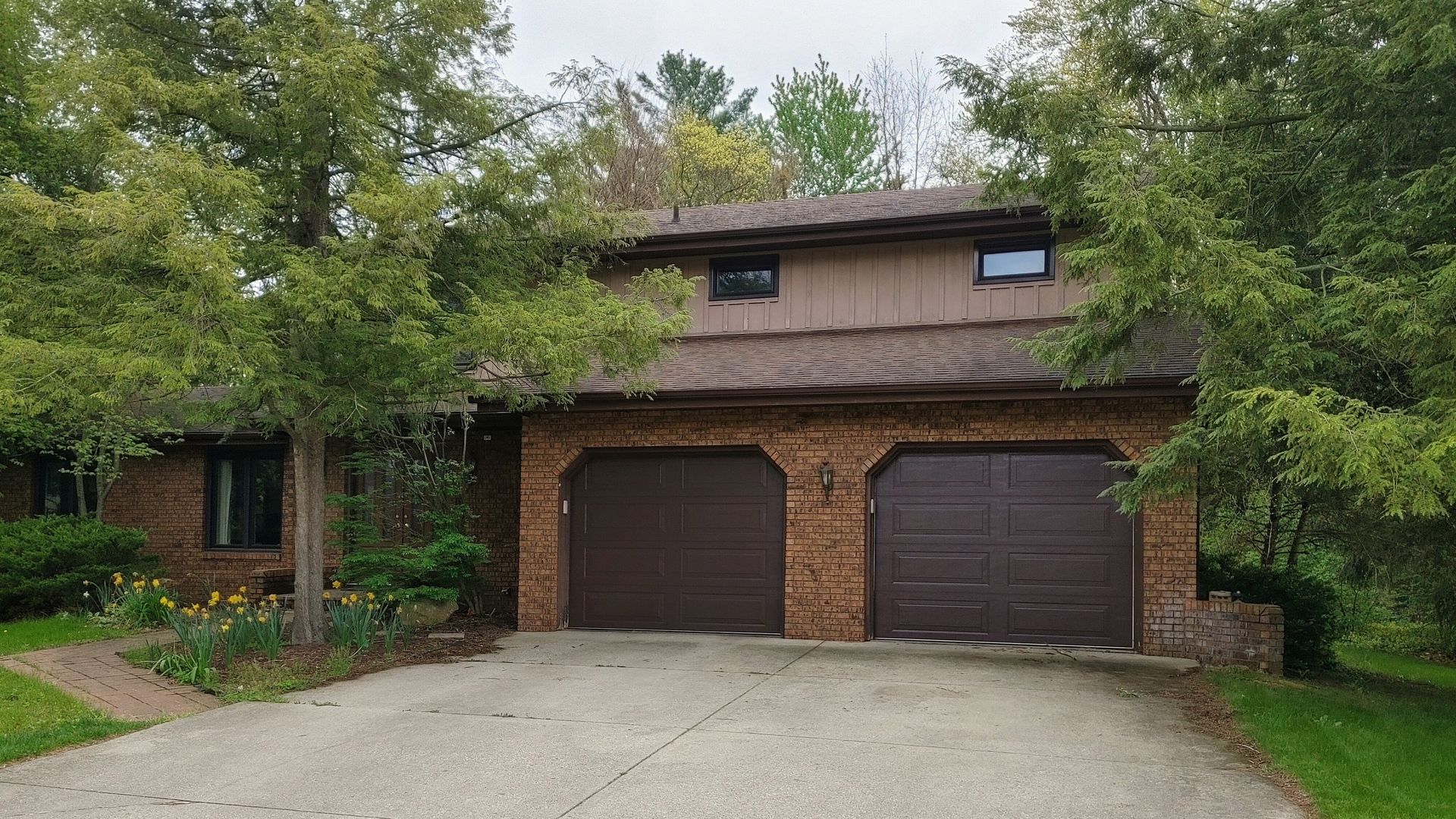 Brick house with two dark garage doors, a gravel driveway, and trees overgrown around the front.