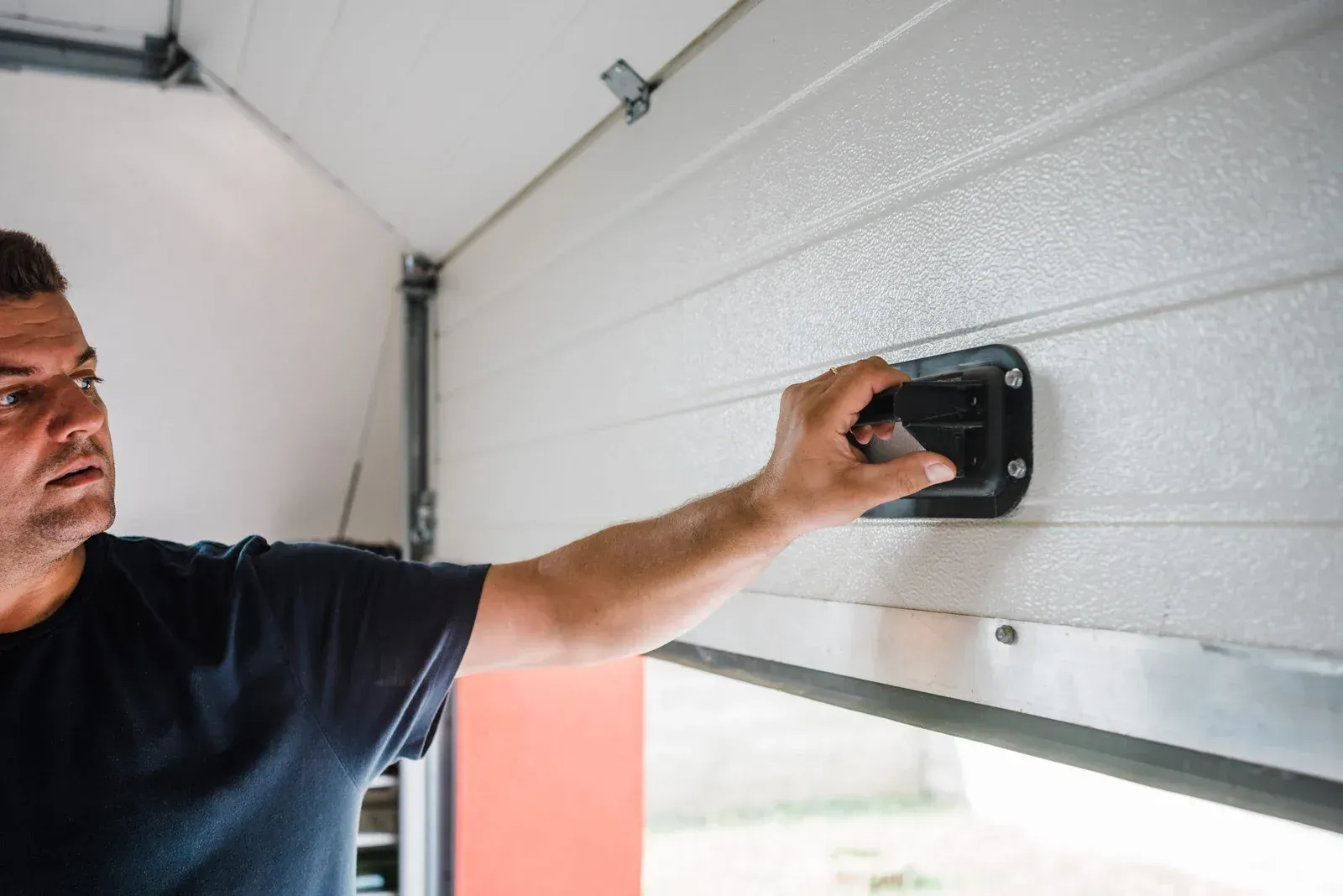 Man inspecting a garage door, possibly fixing it. Indoor setting, white door, black handle.
