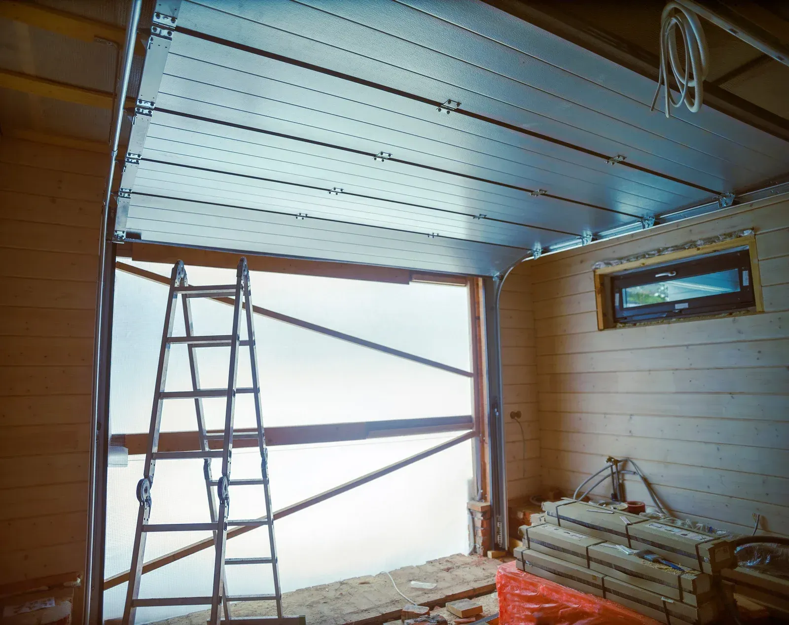 Interior view of a garage under construction; metal door is open, ladder inside, wood walls.