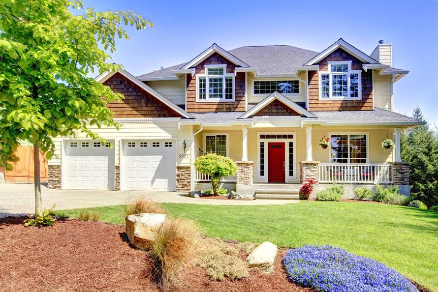 Two-story suburban house with beige siding, red front door, and a two-car garage, framed by a landscaped yard.