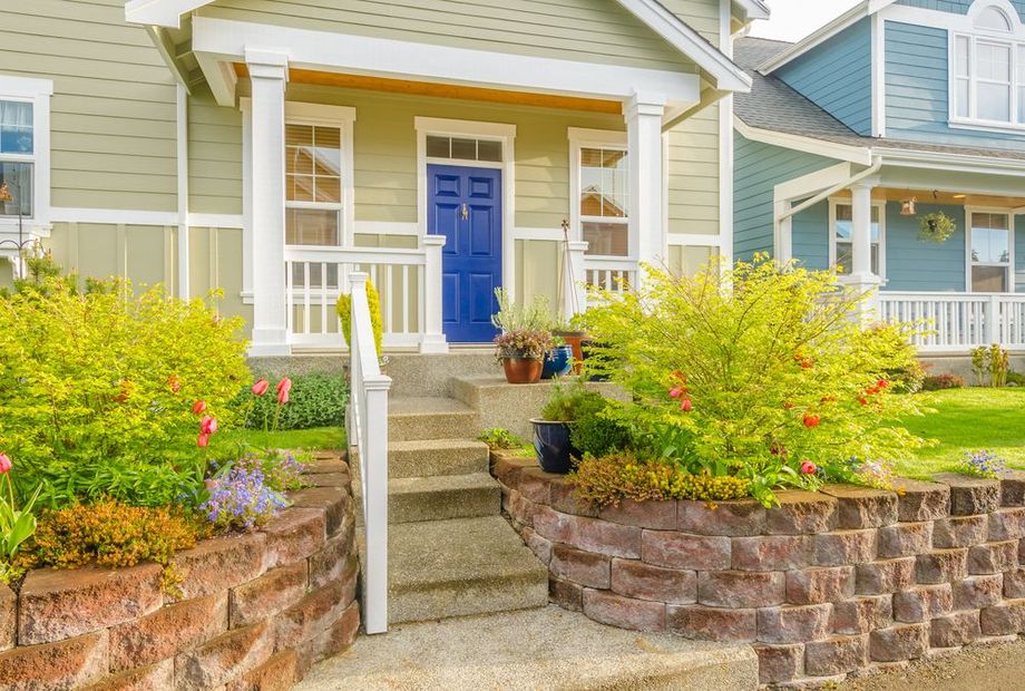 Front porch of a light-green house with blue door, white railings, stone steps, and bright flower beds.