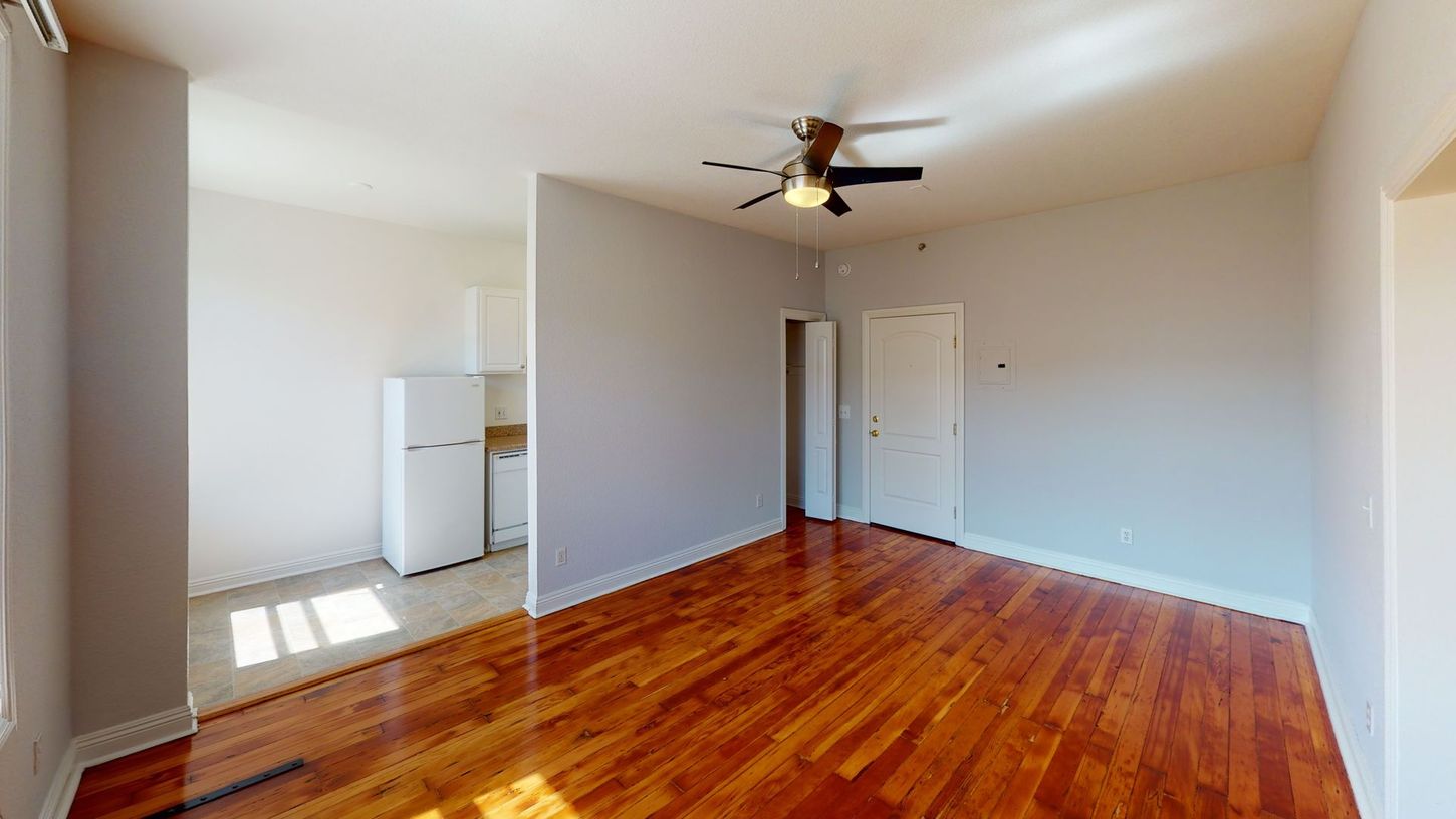 Empty room with hardwood floors, kitchen area, and ceiling fan.