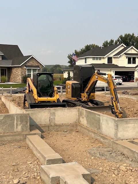 A yellow skid steer and a small excavator sit inside the concrete foundation of a house under construction.