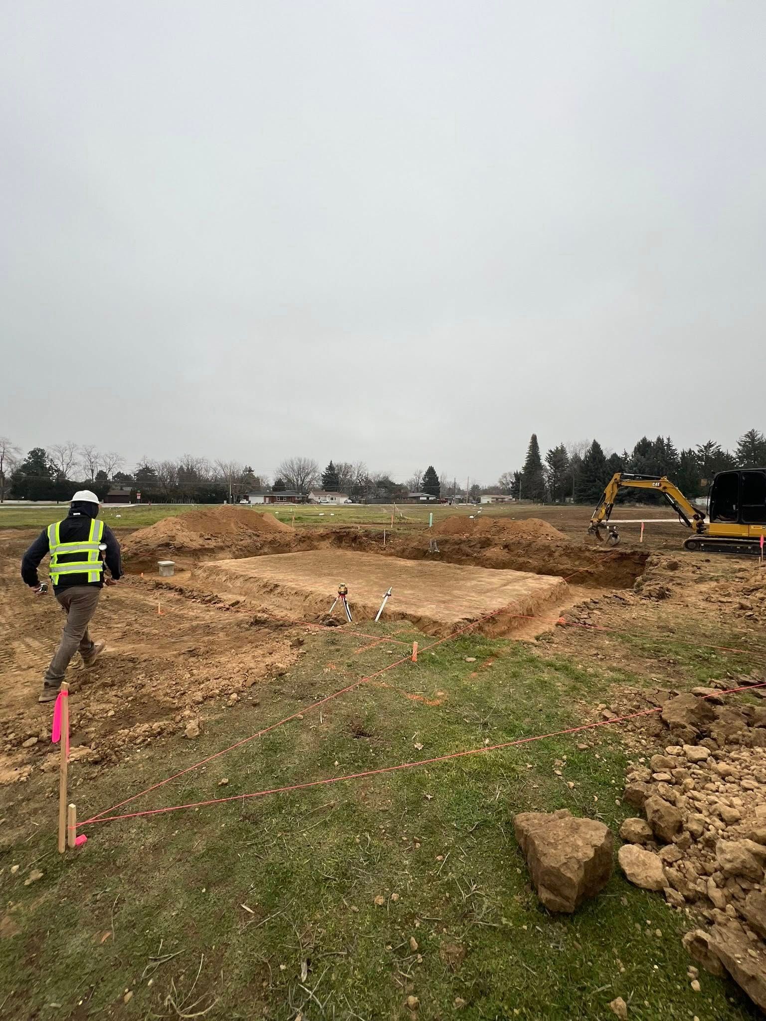 A construction worker in a high-visibility vest walks near a rectangular excavation site with an excavator nearby.