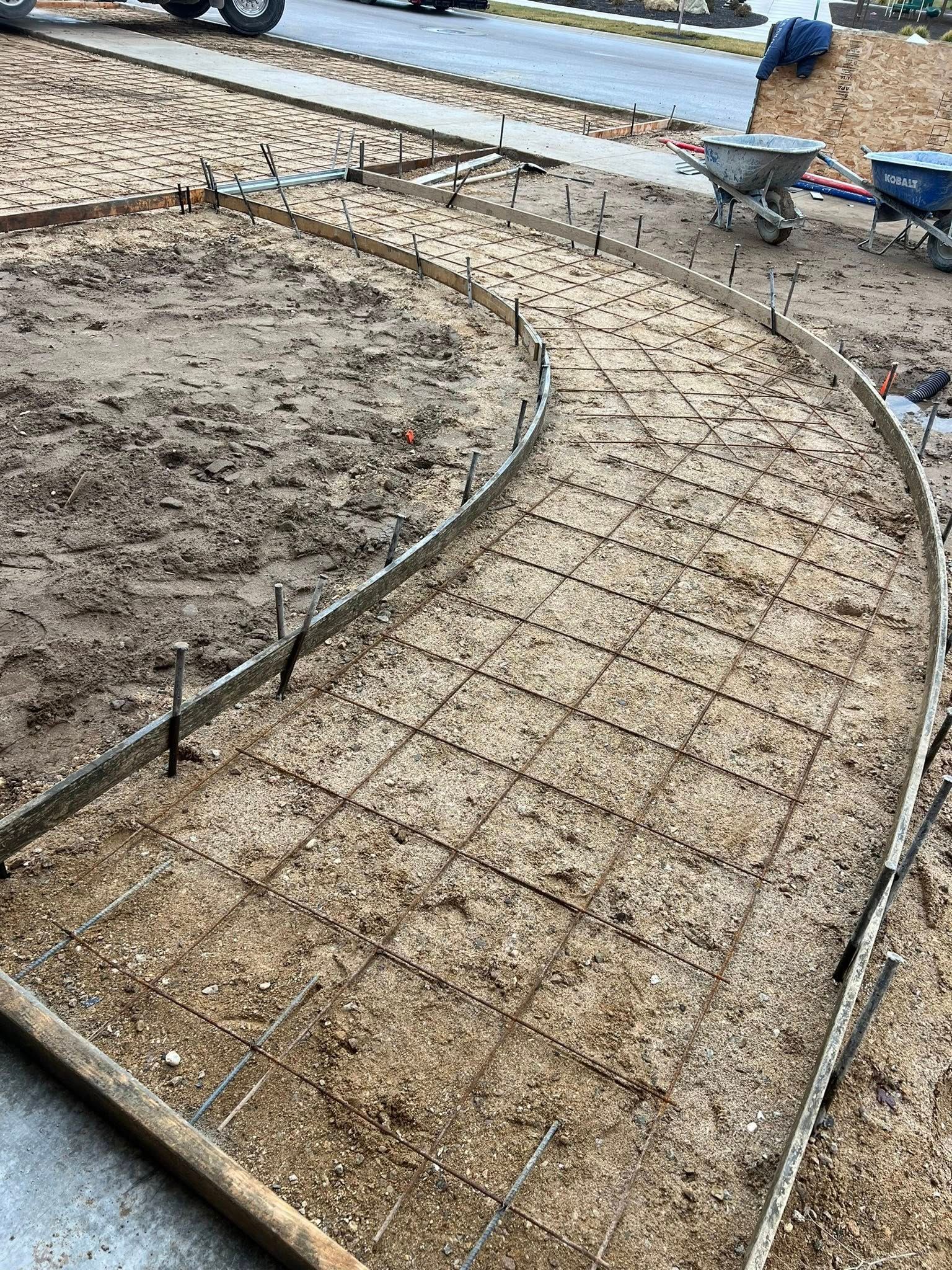 A curving concrete walkway frame prepped with wire mesh and wooden forms, set in a dirt yard with a street in the back.