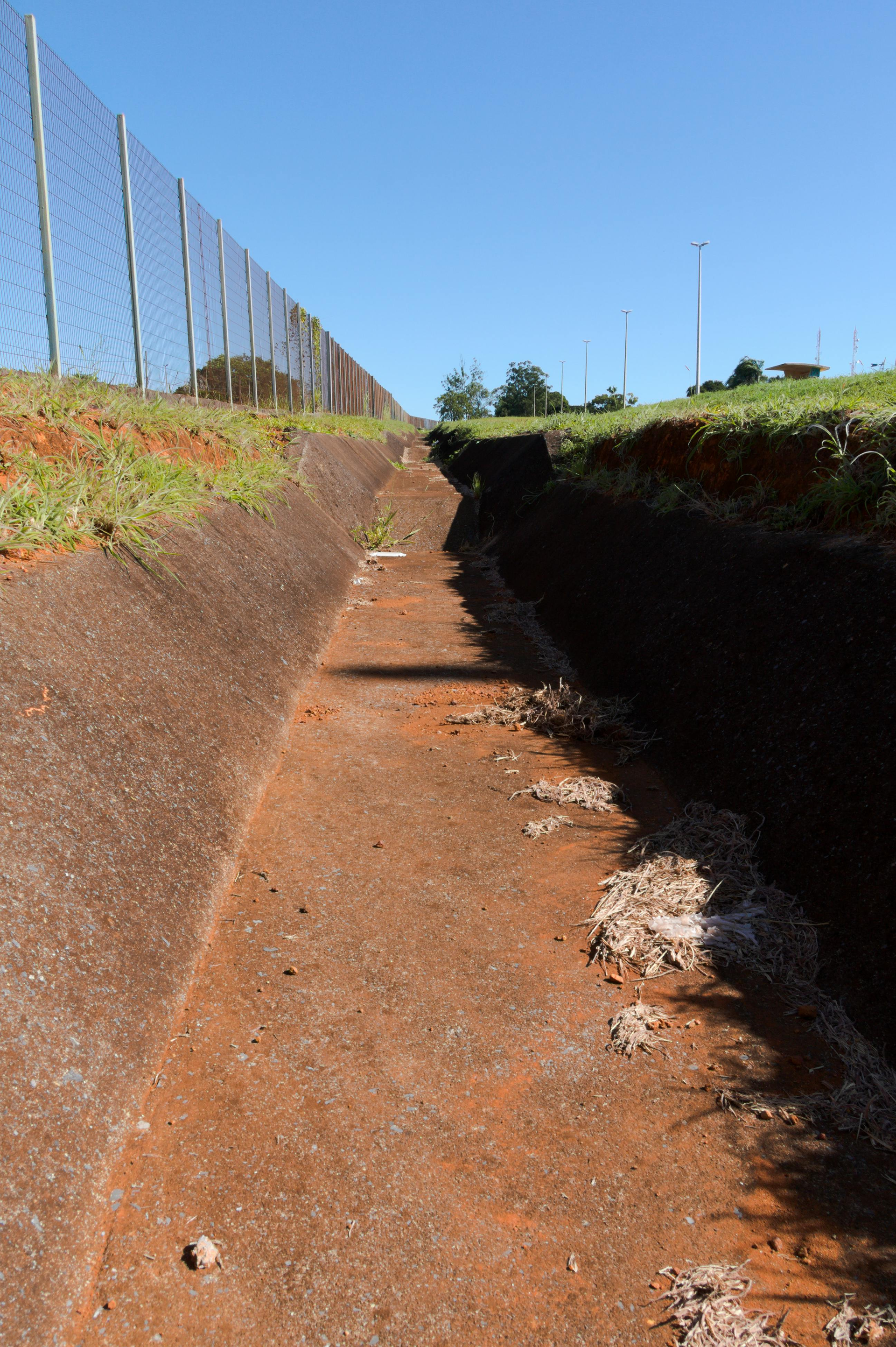 A narrow drainage ditch with a concrete side runs alongside a chain-link fence under a clear blue sky.