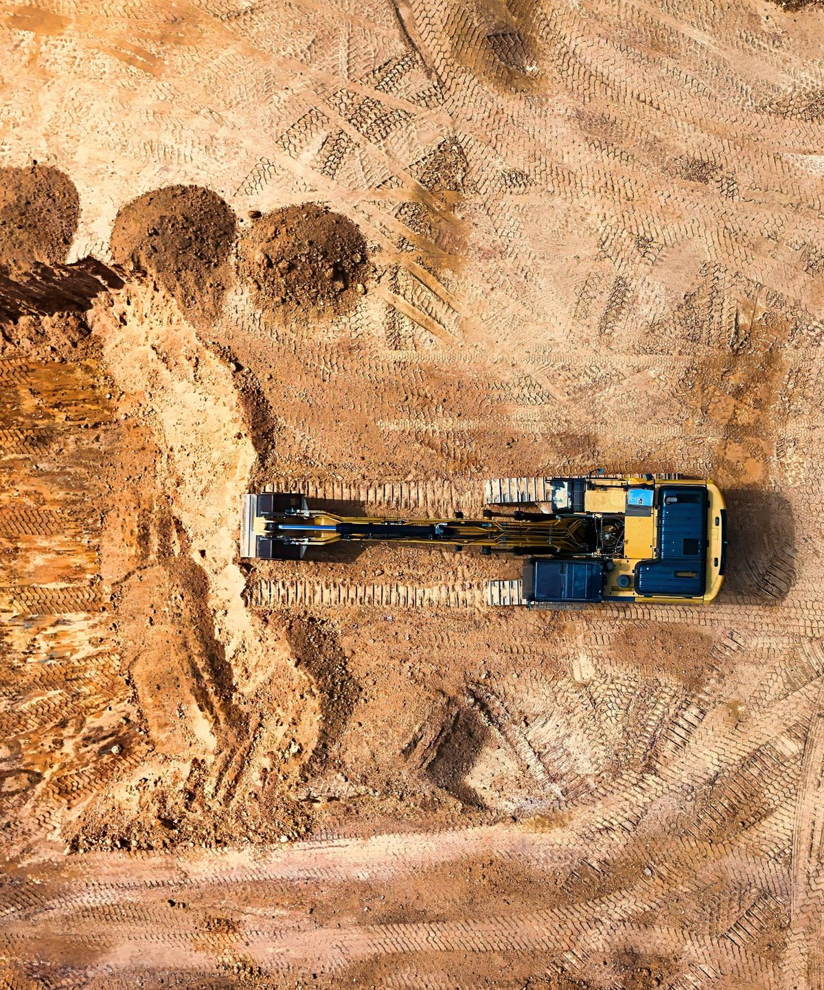 An overhead view of a yellow excavator digging in a large, sandy construction site with piles of excavated earth.