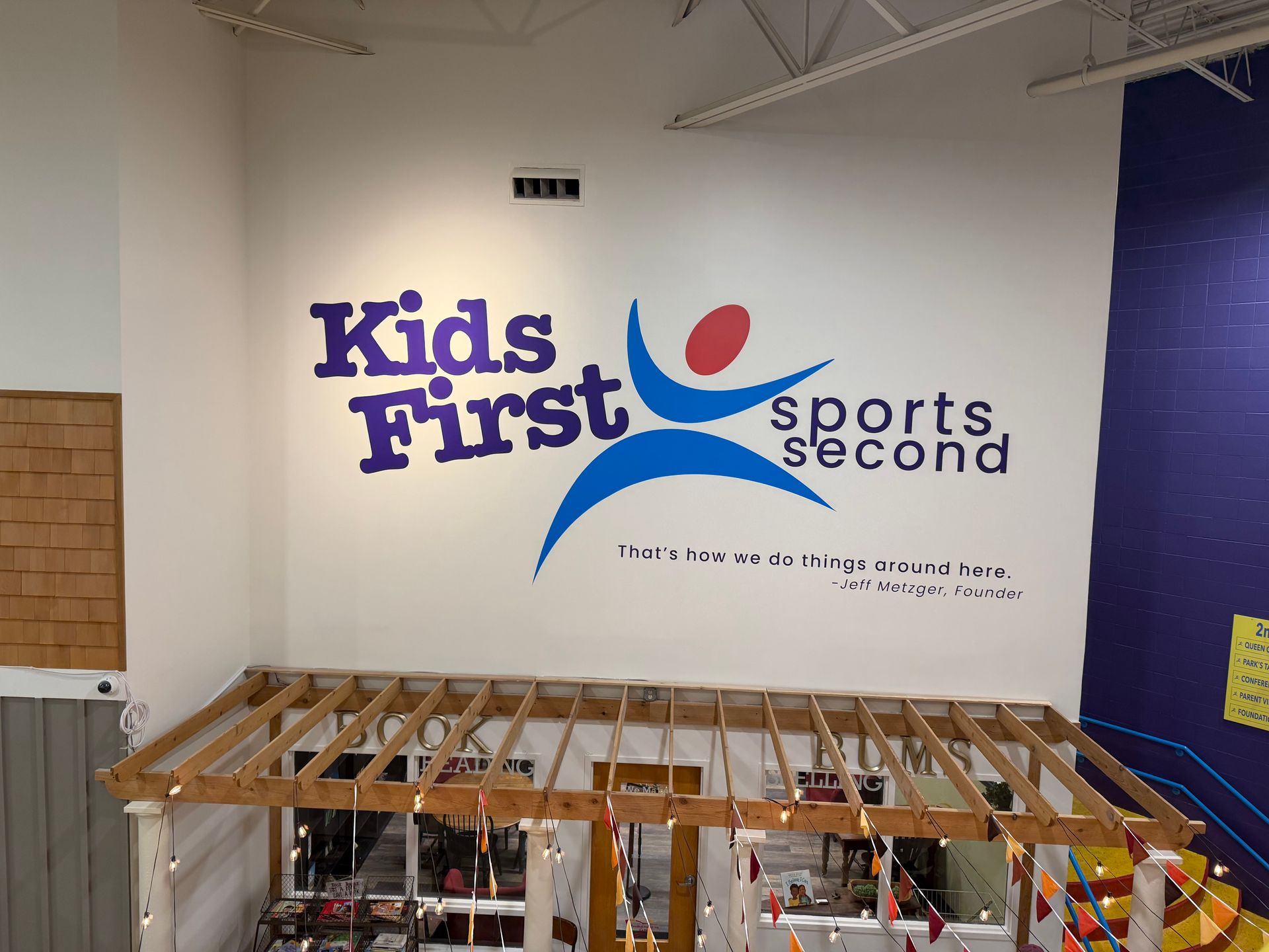 Indoor wall mural reading “Kids First, Sports Second” above a wooden frame structure