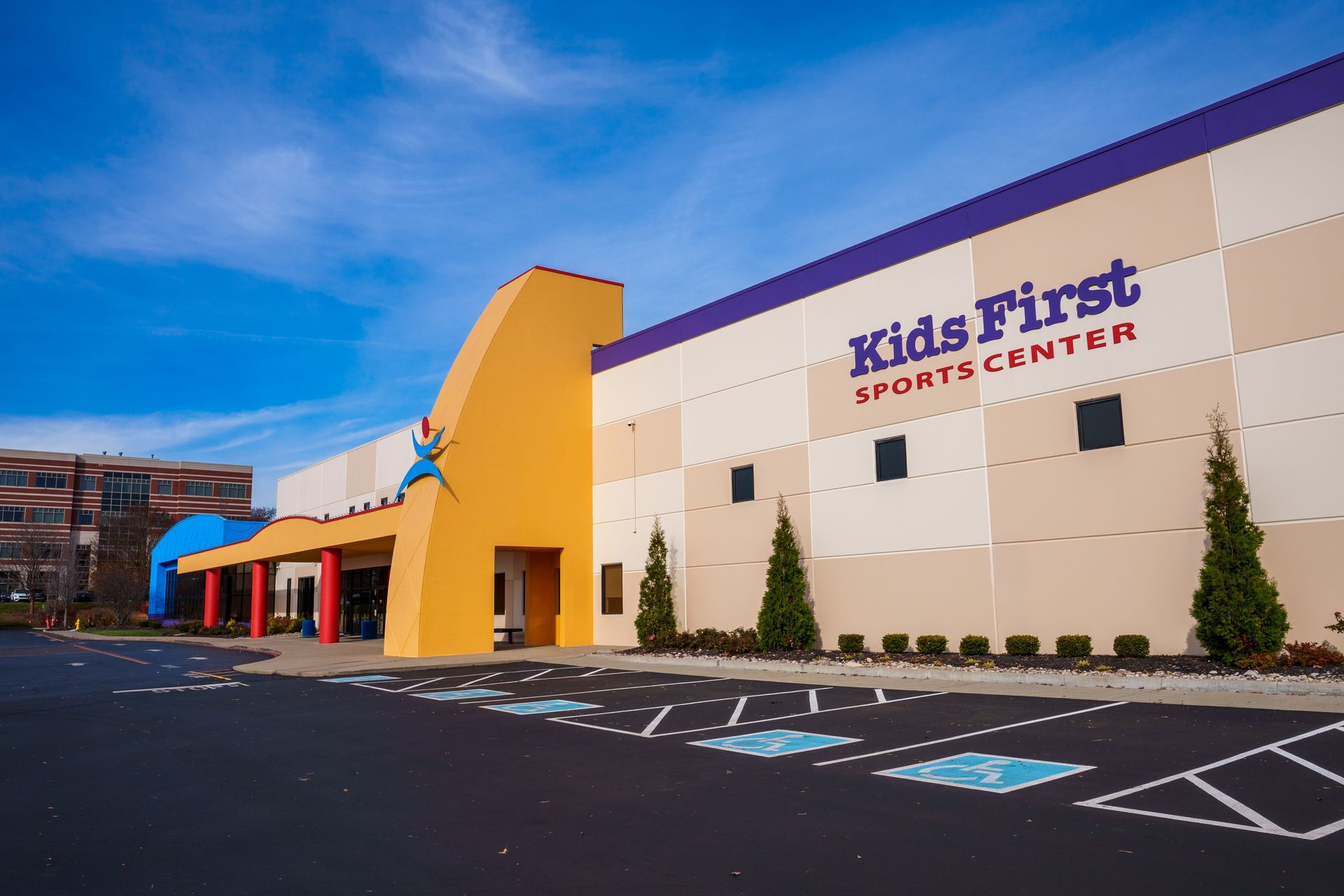 Kids First Sports Center building exterior with beige walls, yellow entrance, and blue awning at dusk