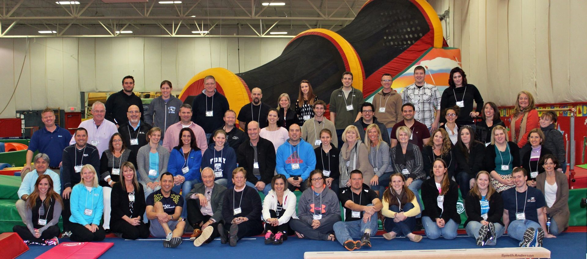 Large group posing indoors in front of a giant black-and-orange tunnel inflatable obstacle course