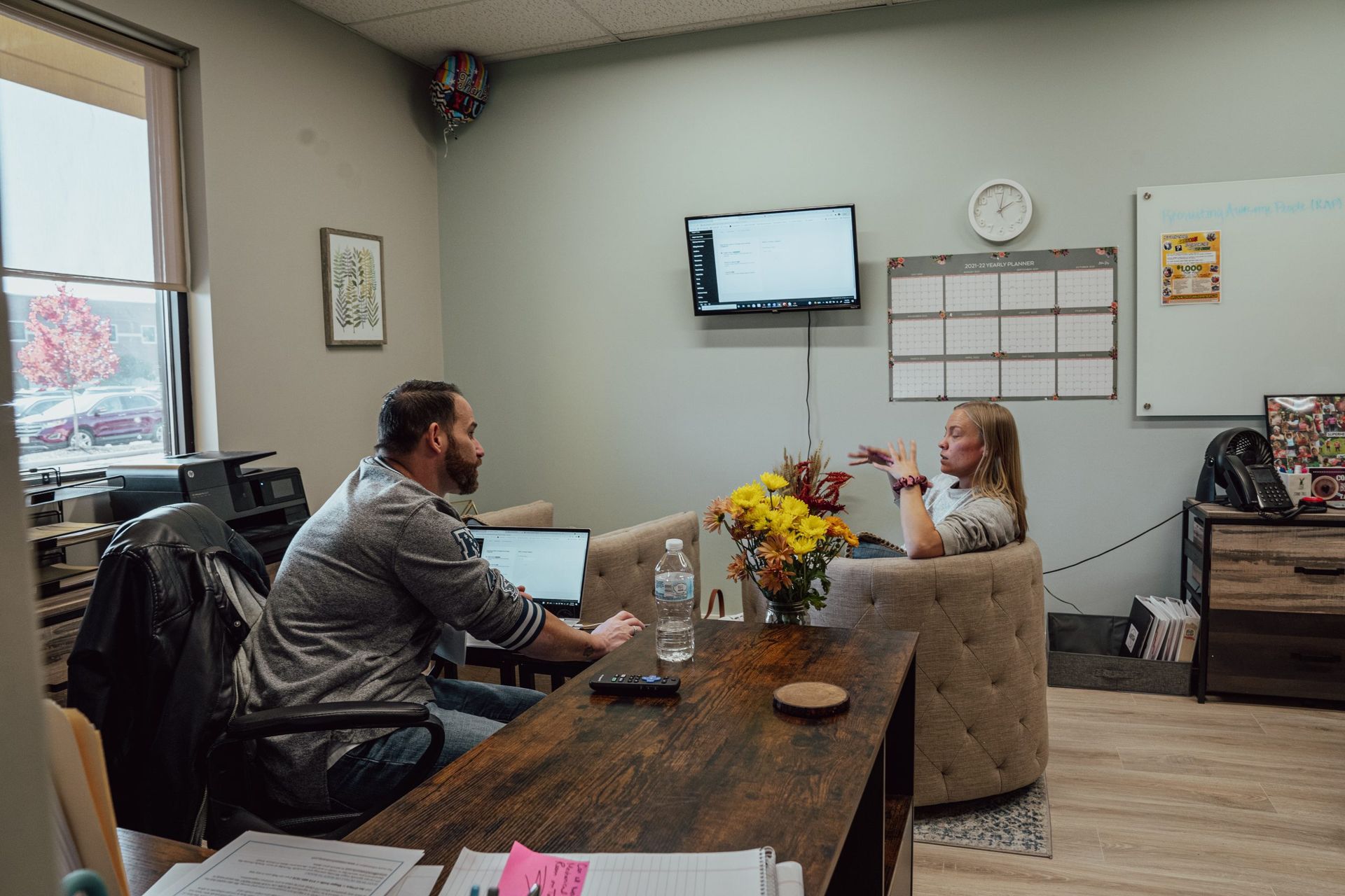 Two people talking across a desk in a small office, with a wall-mounted TV and calendar behind them.