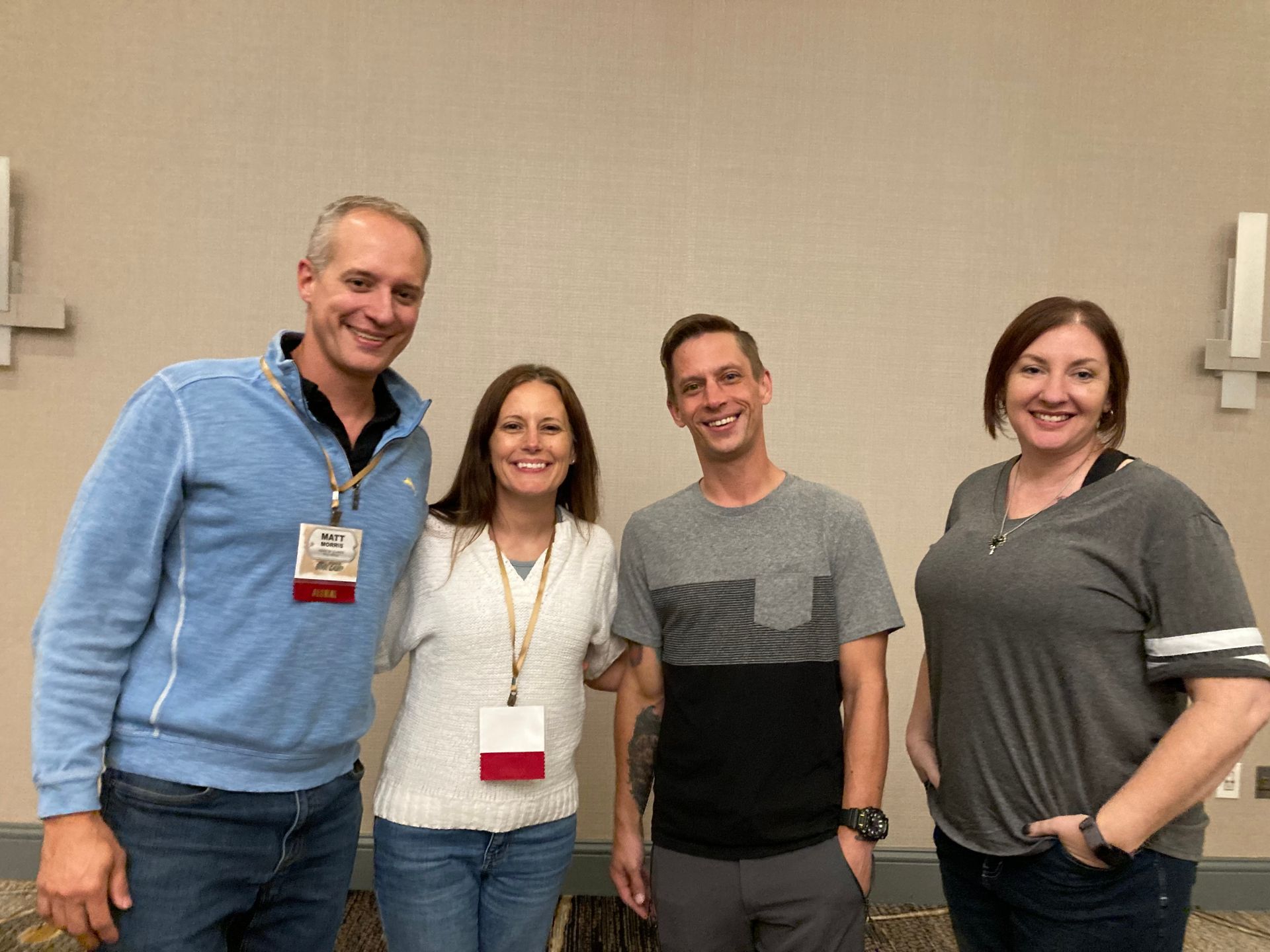 Four people smiling and posing indoors with lanyards against a beige wall