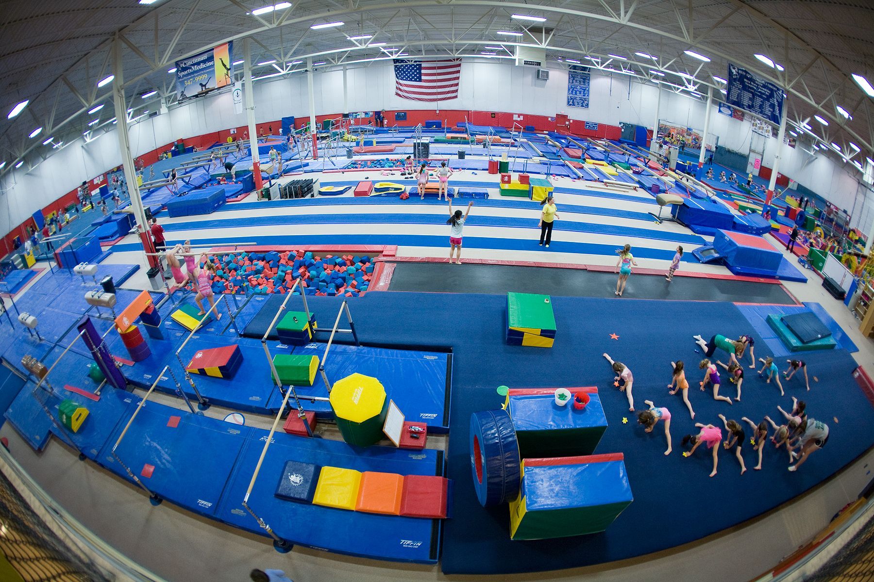Large indoor gymnastics gym with blue mats, colorful equipment, and athletes practicing routines