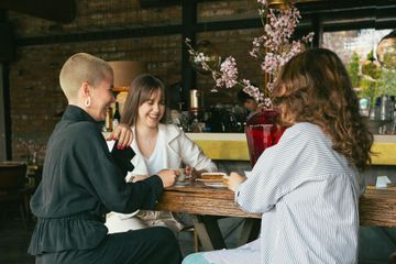 Three people chatting at a café table, smiling with drinks and flowers in the background.