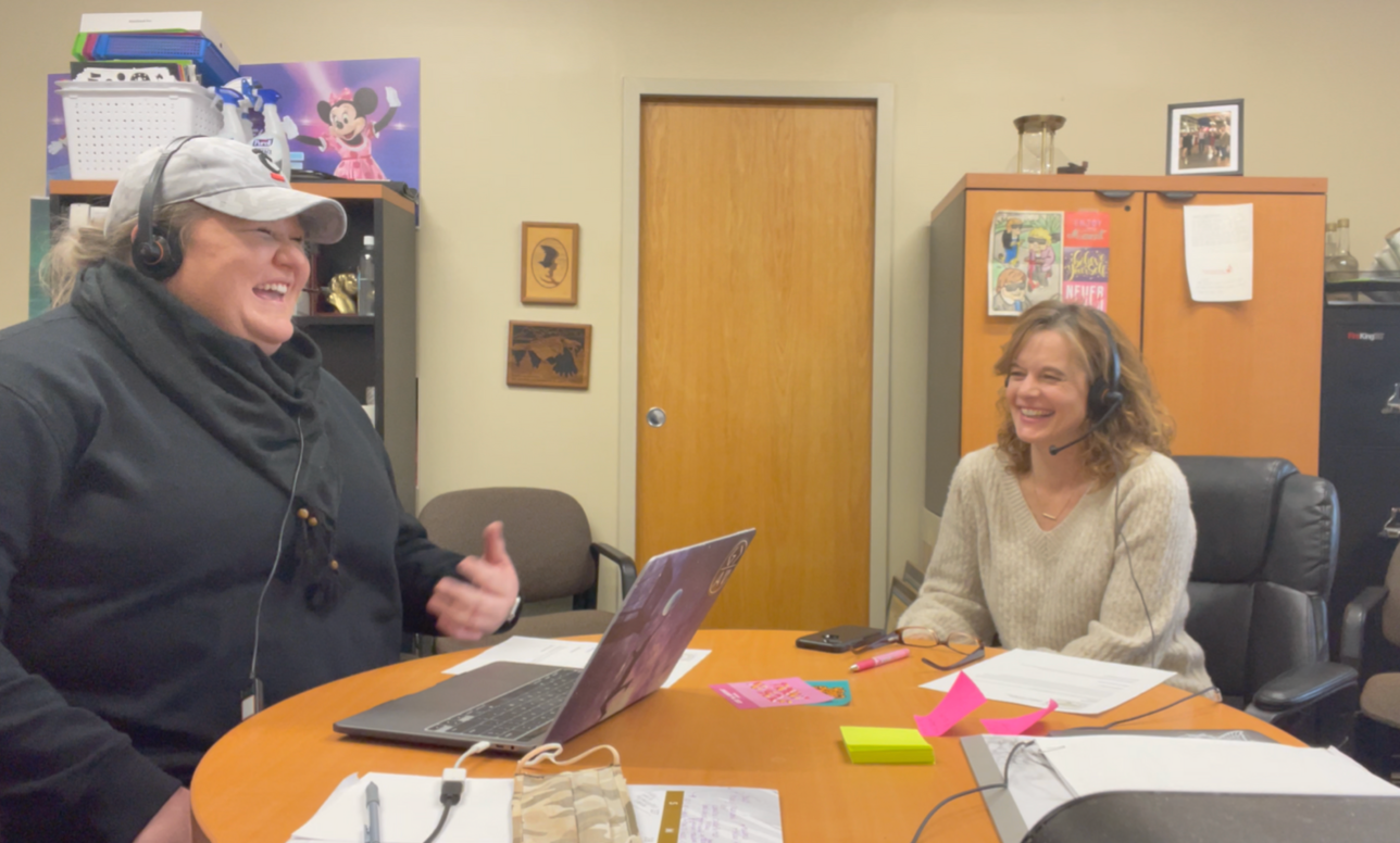Two people smiling in an office, seated at a table with a laptop and papers.