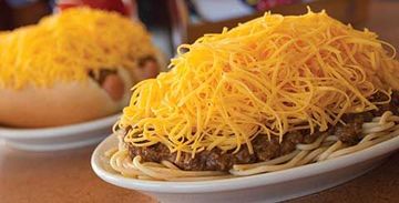 Two hot dogs topped with shredded cheddar on white plates, with one in the foreground and one blurred behind.