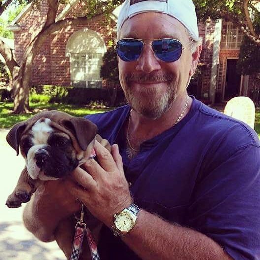 Man in sunglasses holding a brown and white bulldog puppy. Outdoors, brick house in background.