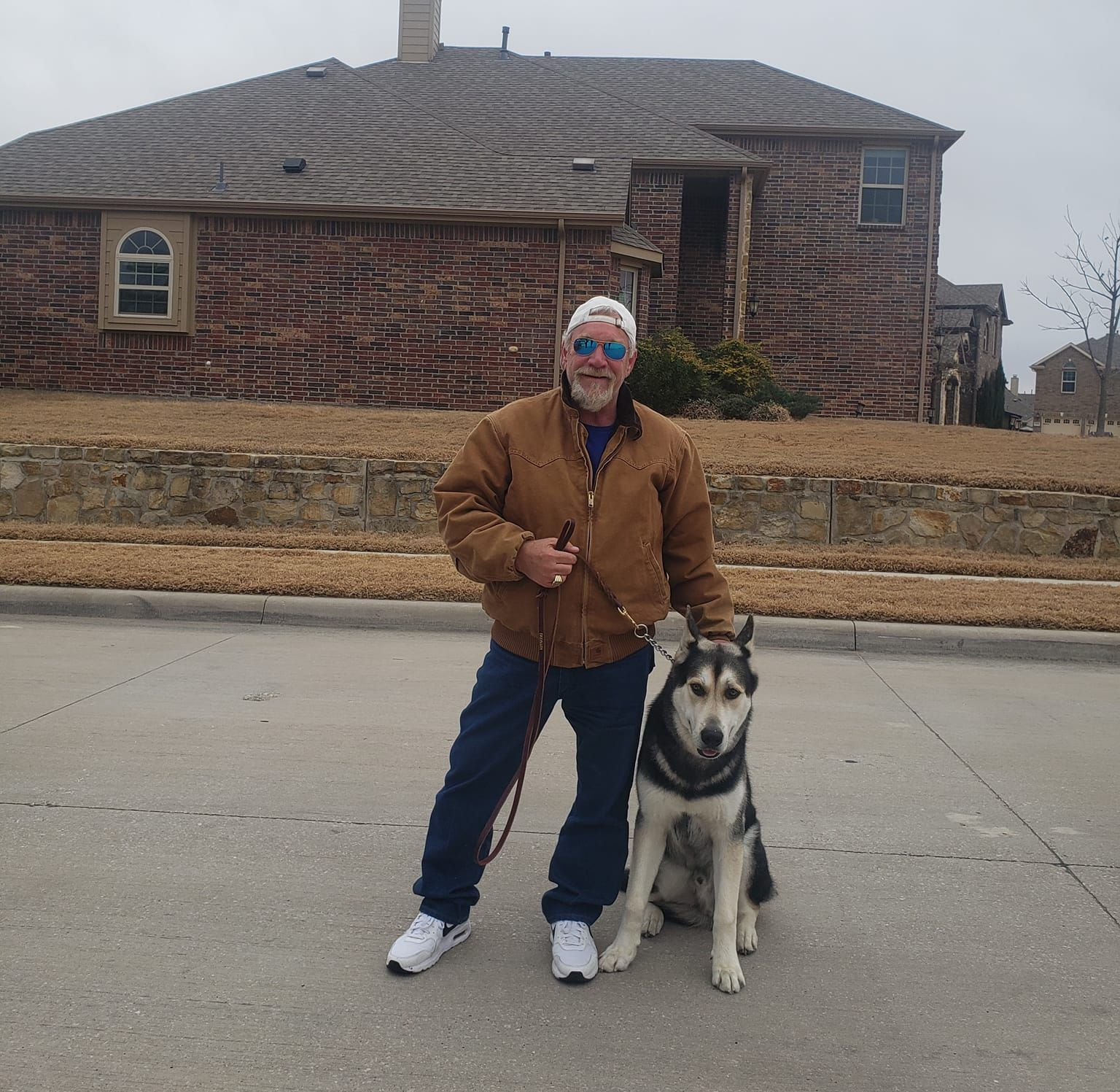 Man in a brown jacket and blue jeans, standing with a husky dog on a street in front of a brick house.