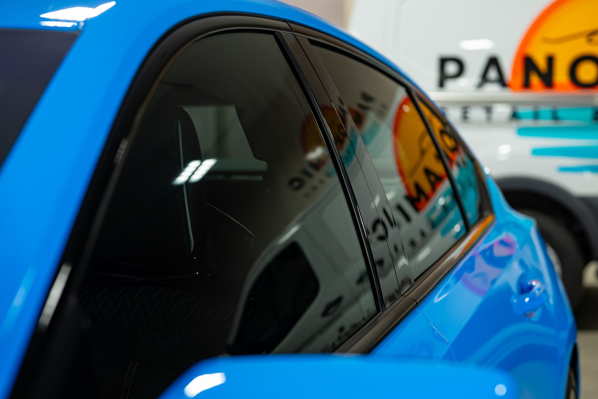 A close-up of a bright blue car’s windows reflecting the logo of a detailing business on a nearby white van.