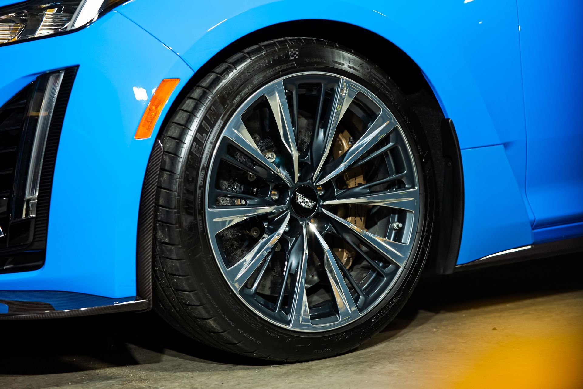 A close-up view of a bright blue car's front tire and chrome multi-spoke wheel against a dark background.