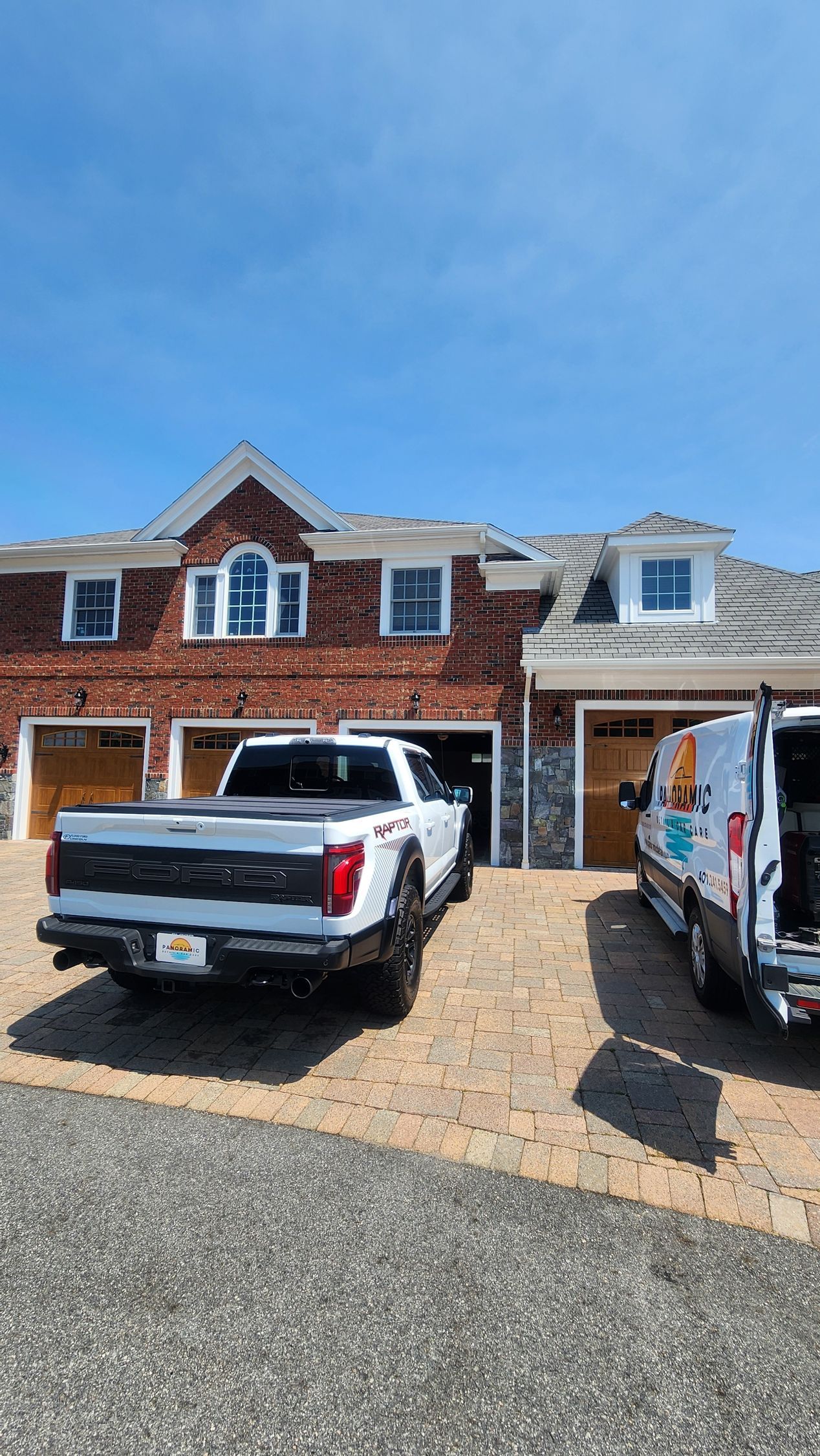 White pickup truck and van parked in front of a brick house with open garage doors on a sunny day.