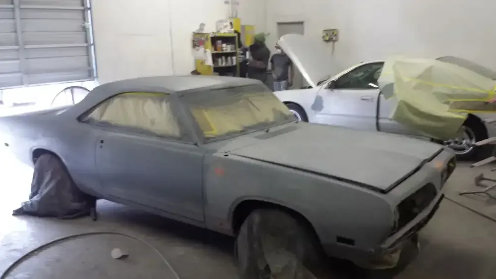 A grey, primer-coated classic car sits in a repair shop with another masked-off car in the background.
