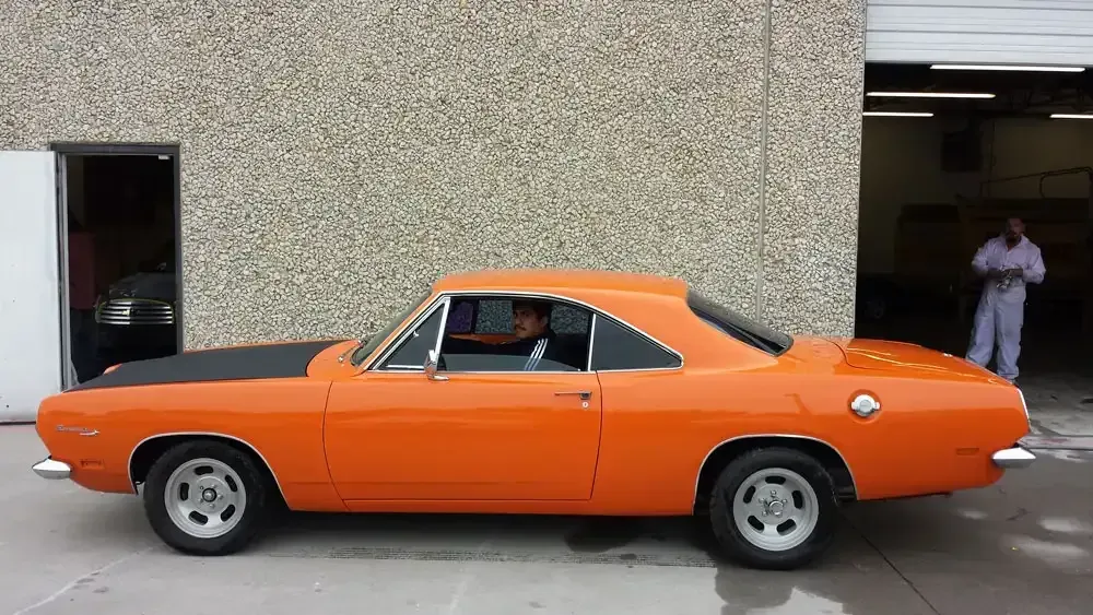 A bright orange classic coupe parked outside a workshop, with a person sitting in the driver's seat.