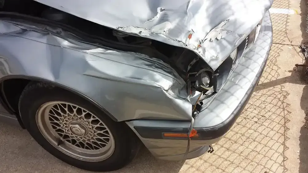 A silver car with significant body damage to the front hood, fender, and bumper, parked near a chain-link fence.