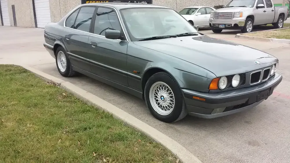 A dark green BMW 5 Series sedan parked on an asphalt lot with a curb and grass in the foreground.