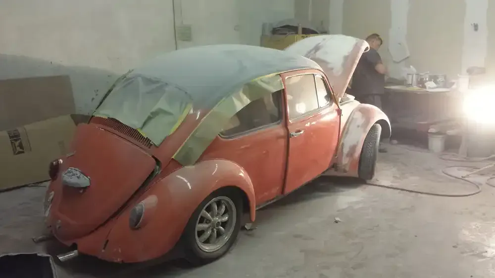An orange Volkswagen Beetle undergoes bodywork and masking in a brightly lit automotive workshop.