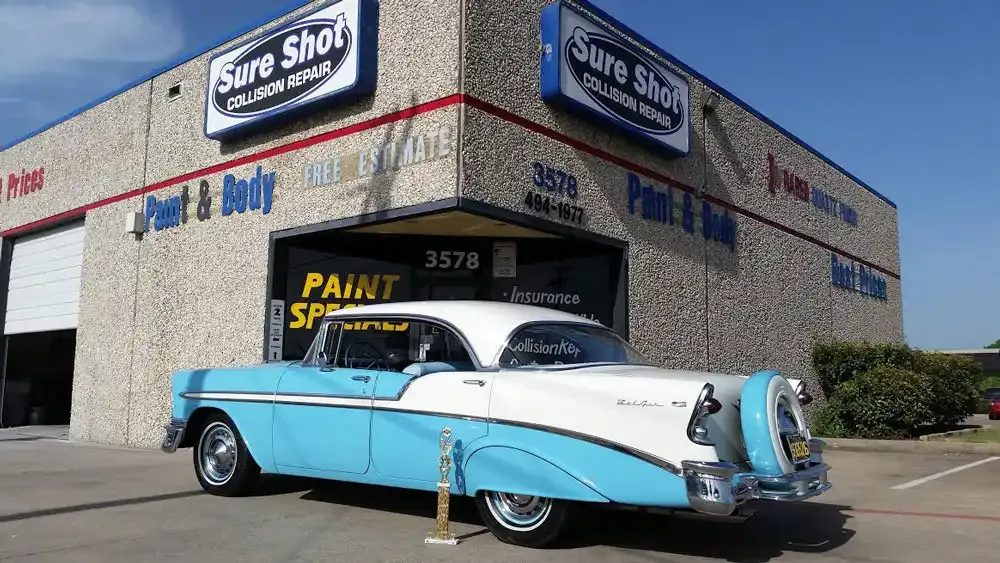 A blue and white classic car parked in front of the Sure Shot Collision Repair shop on a sunny day.